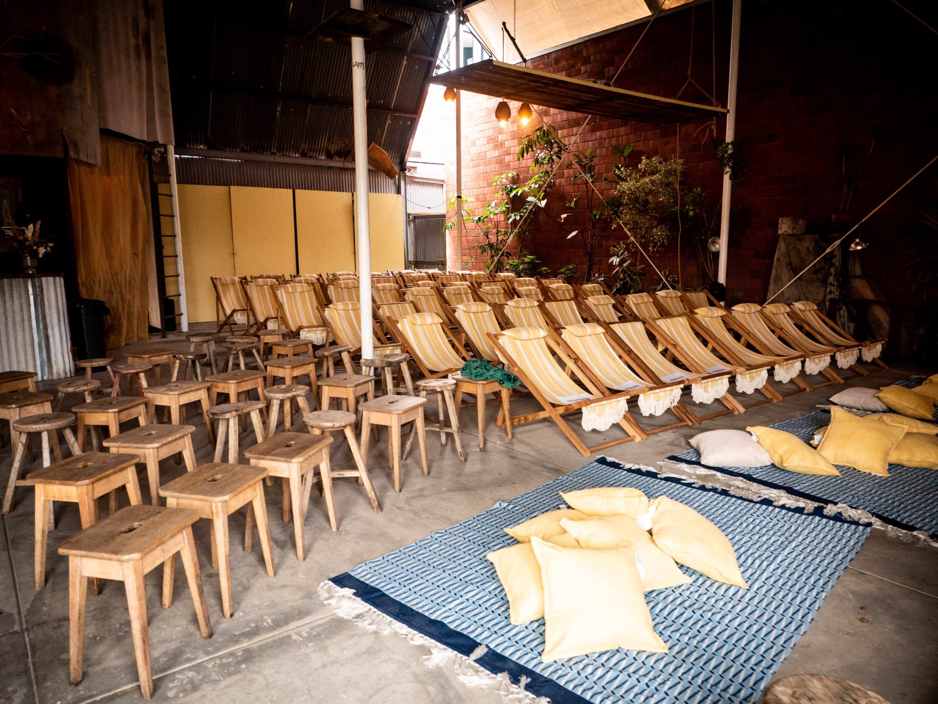 The image shows a spacious indoor venue with rows of wooden chairs, small wooden stools, and floor cushions arranged on a patterned rug.