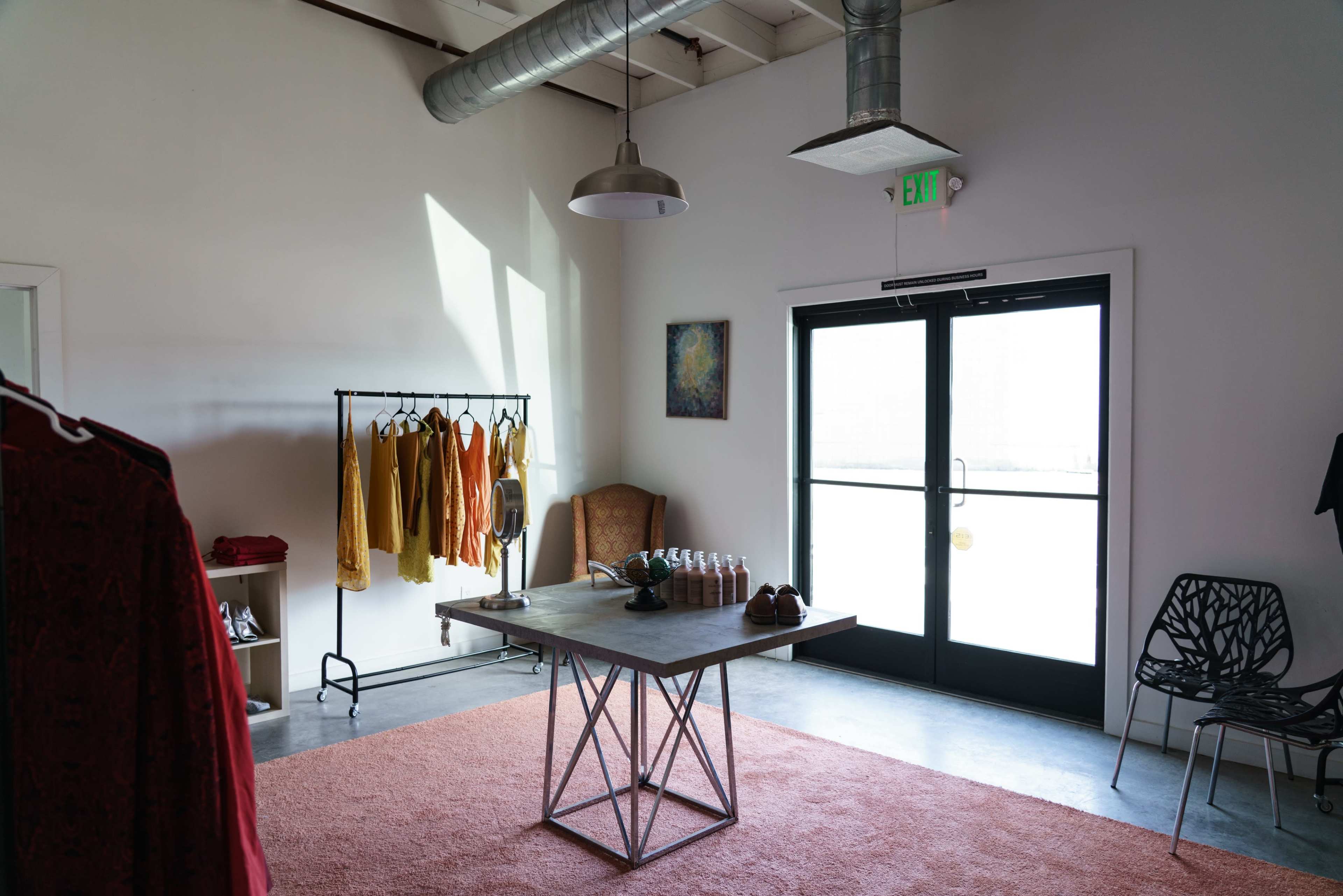 The image shows a minimalist retail space featuring a display rack with clothing, a table with decorative items, and a large window with a door leading outside.