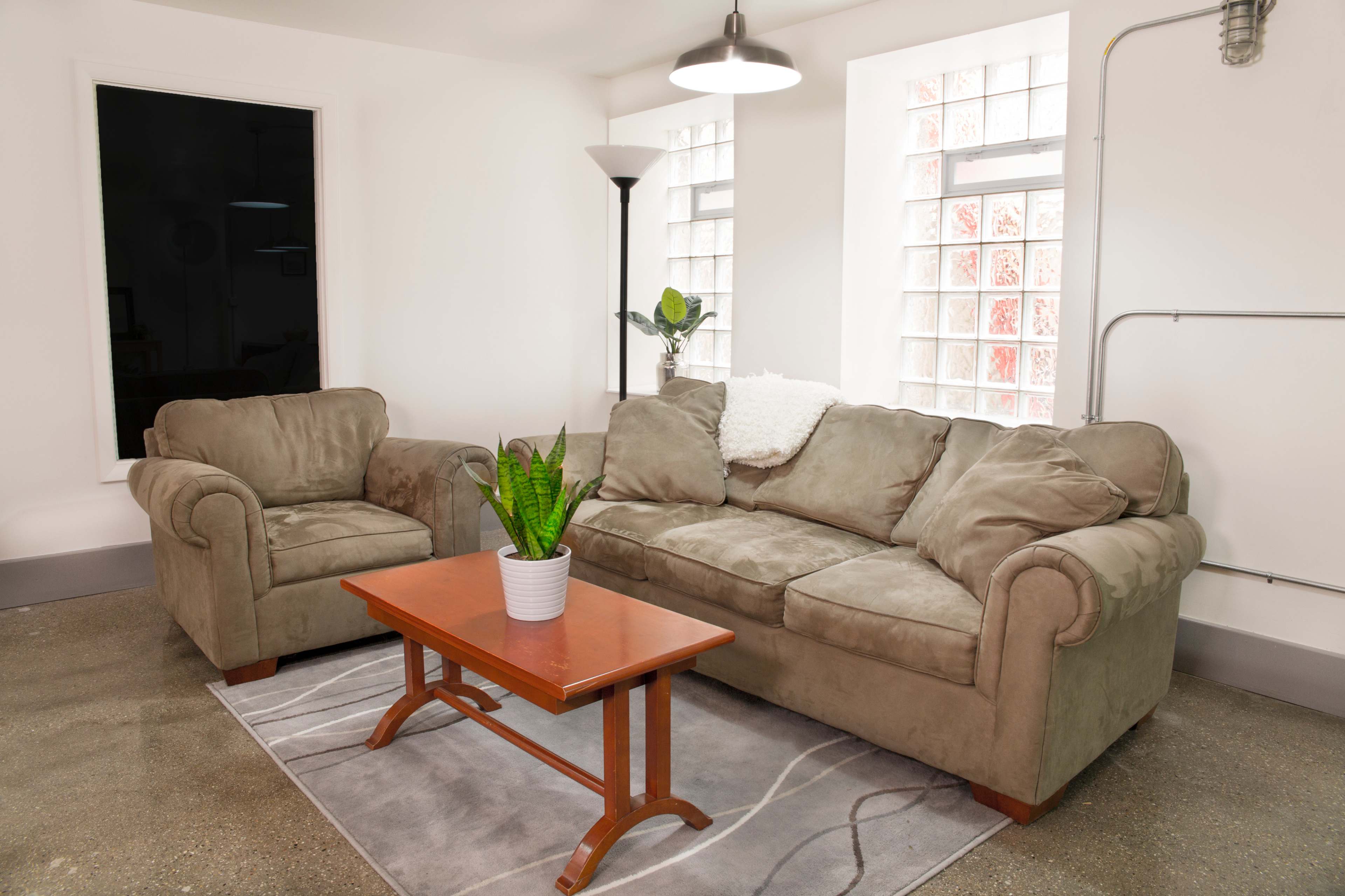 A living room features a beige couch, an armchair, a wooden coffee table, and large windows with glass block designs.