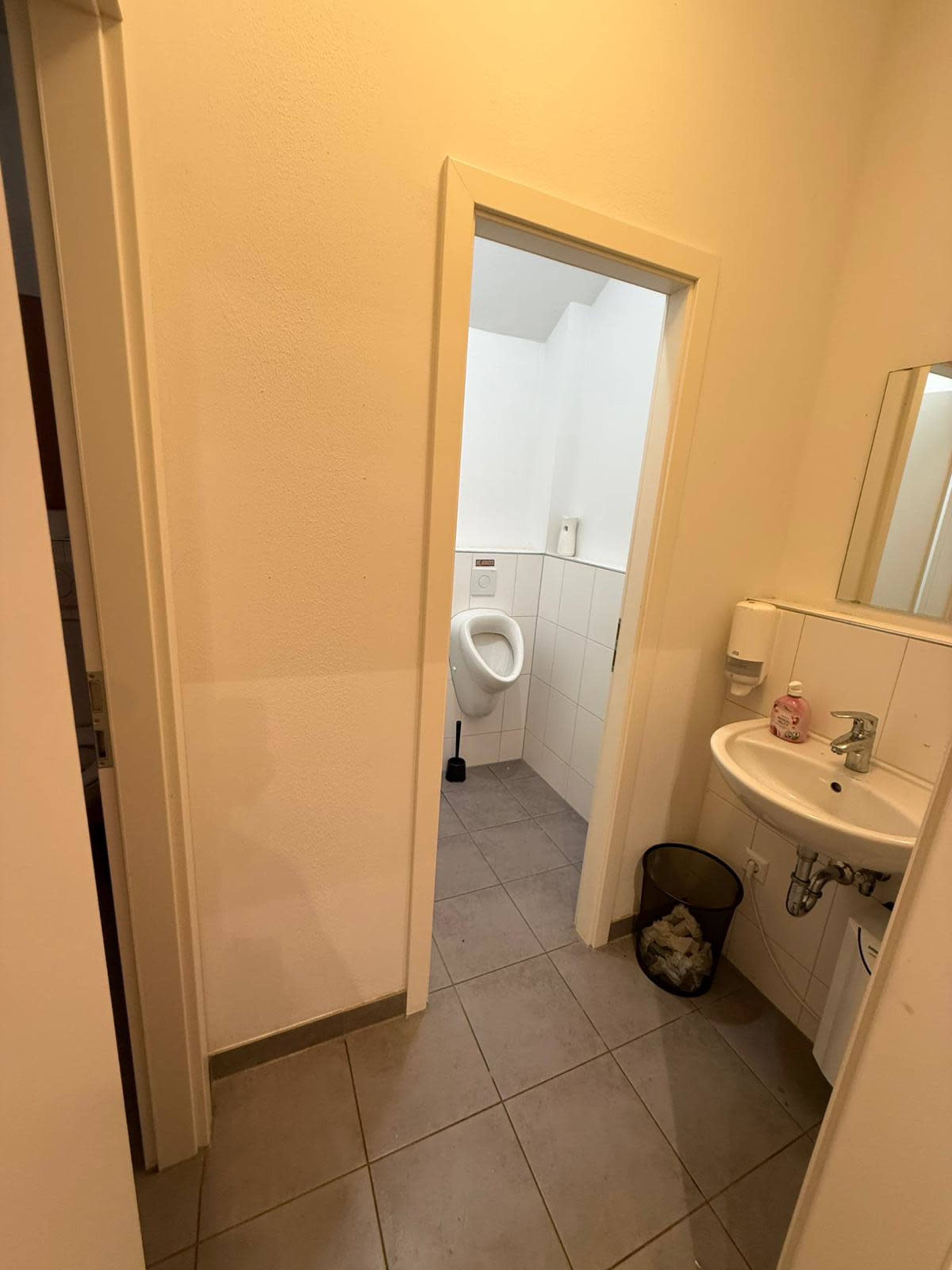The image shows a small bathroom hallway with a urinal on the left, a sink on the right, and a waste bin on the floor.
