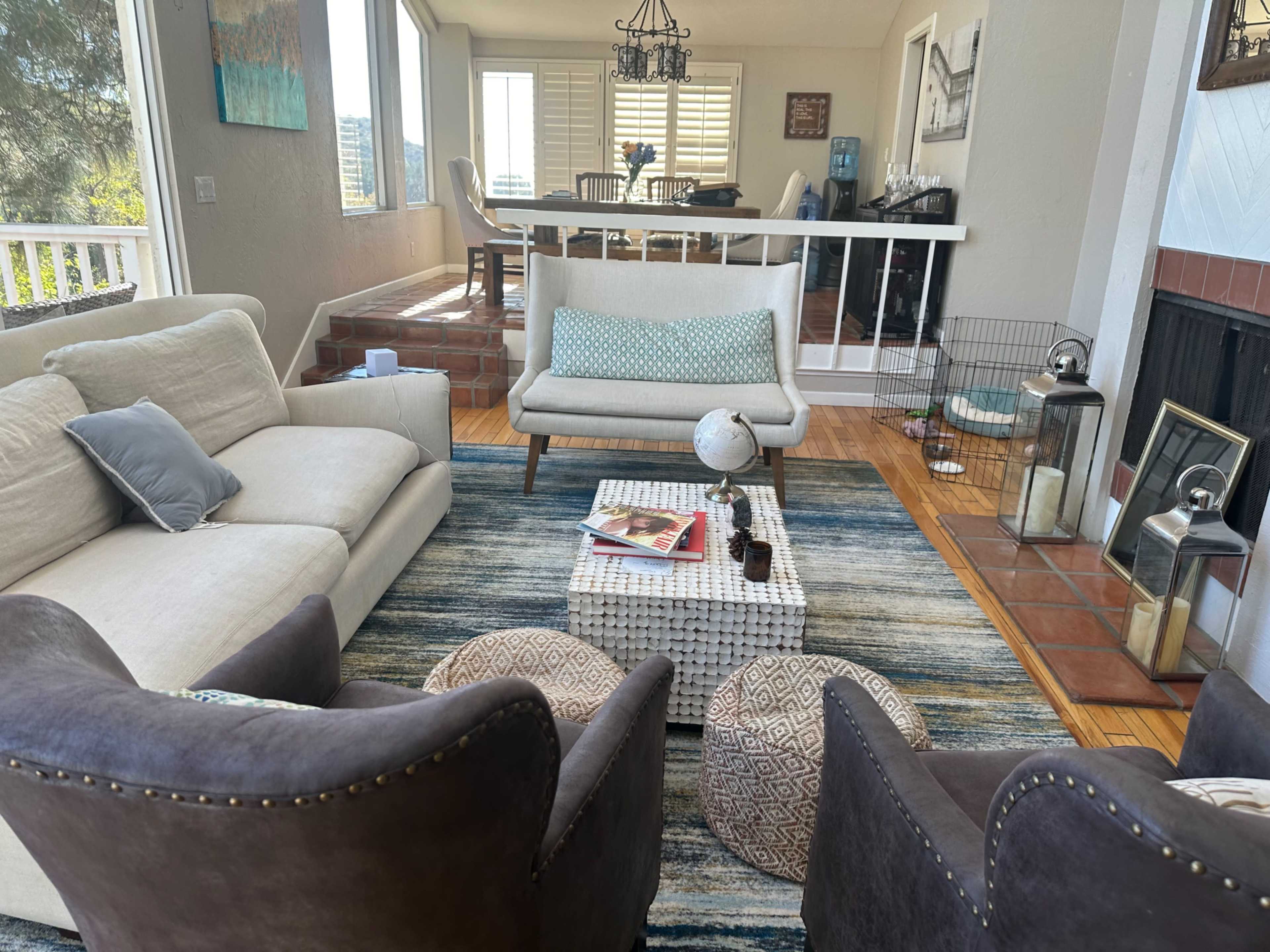 The image shows a living room with a light-colored sofa, two dark armchairs, and a coffee table on a patterned rug, surrounded by large windows and a dining area in the background.