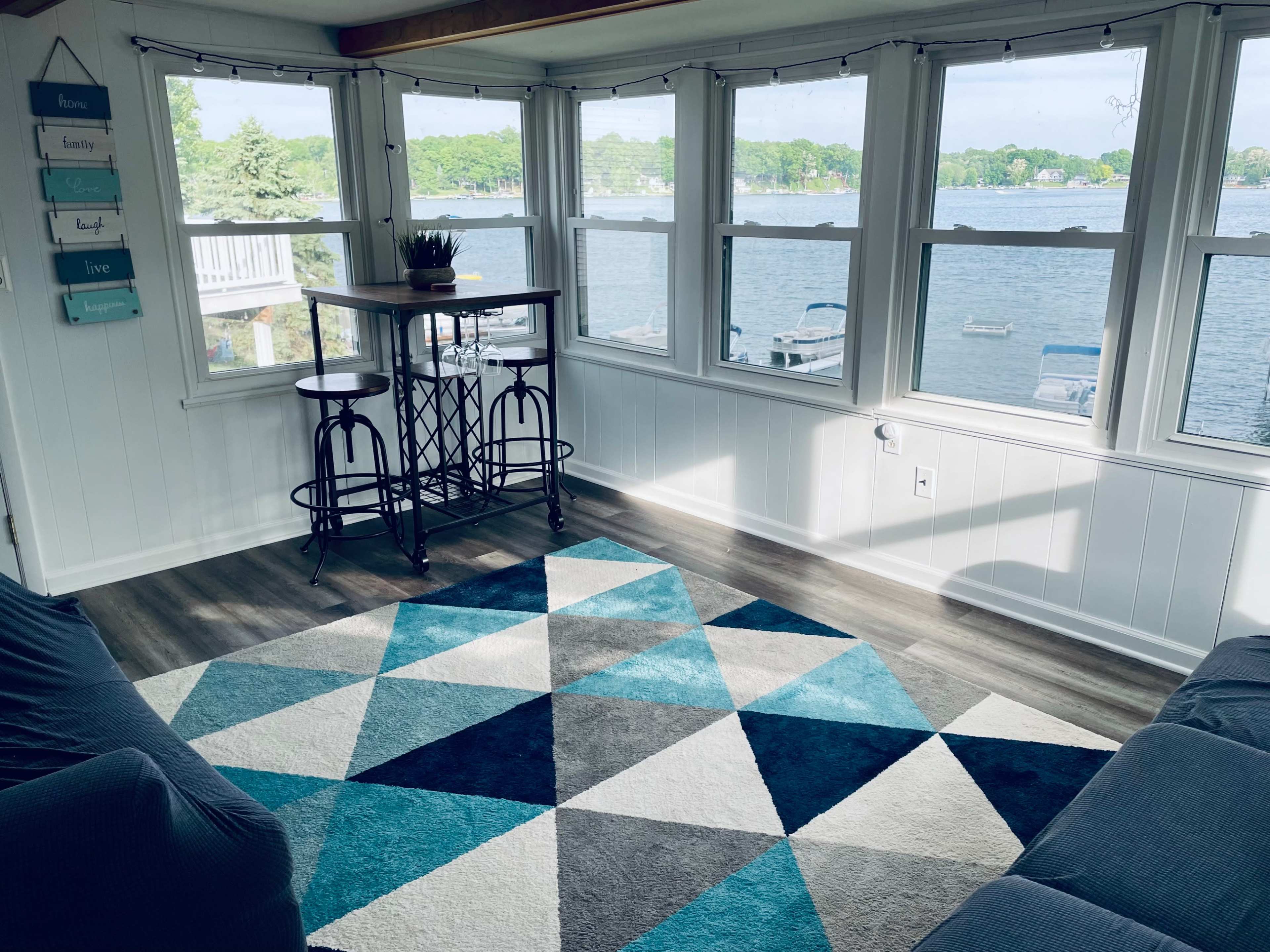 A cozy sunlit room with large windows overlooking a body of water, featuring a geometric-patterned rug and a small bar table with chairs.