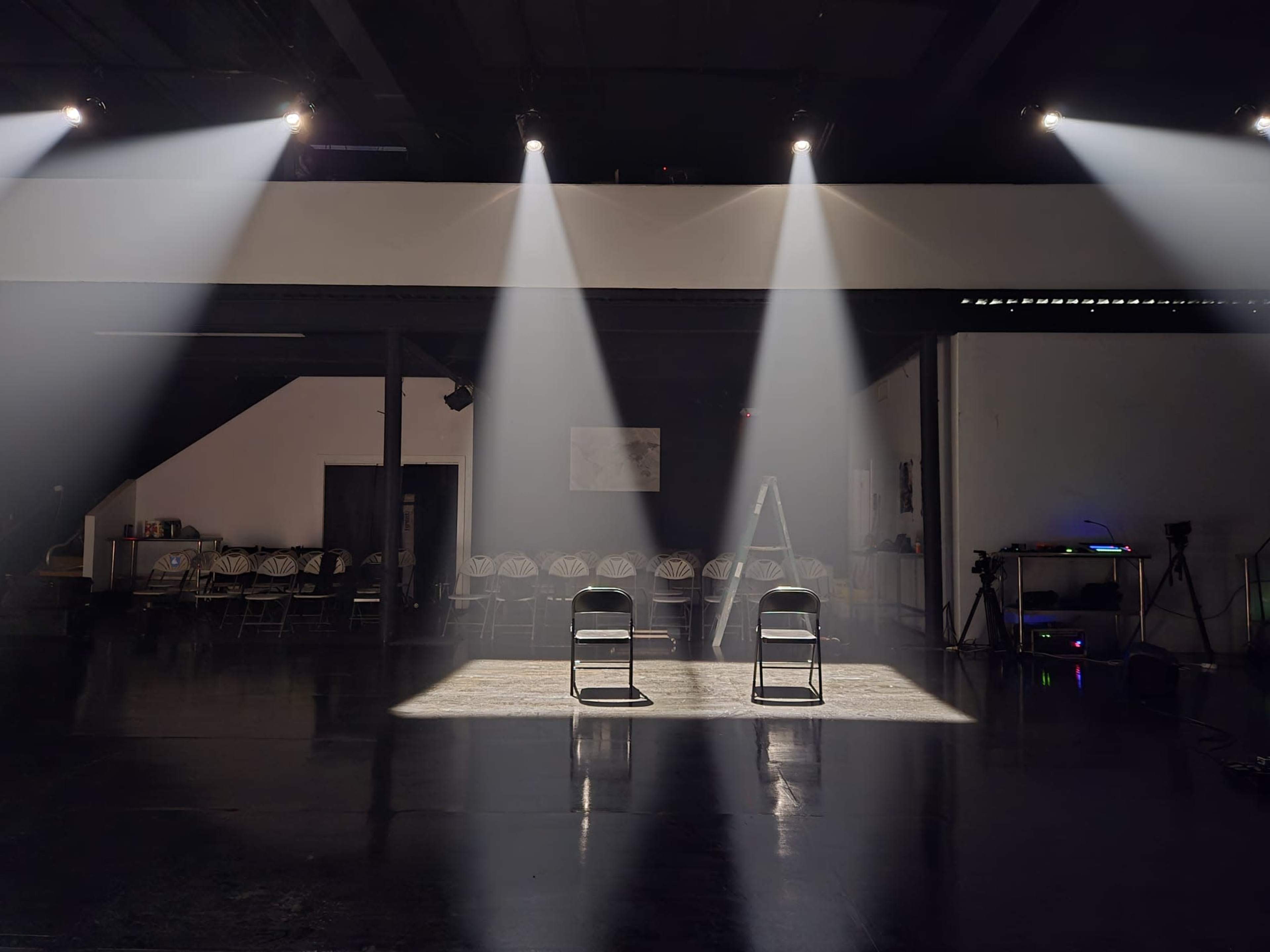 Two empty chairs are illuminated by beams of light on a dark stage, surrounded by rows of empty folding chairs and a ladder.