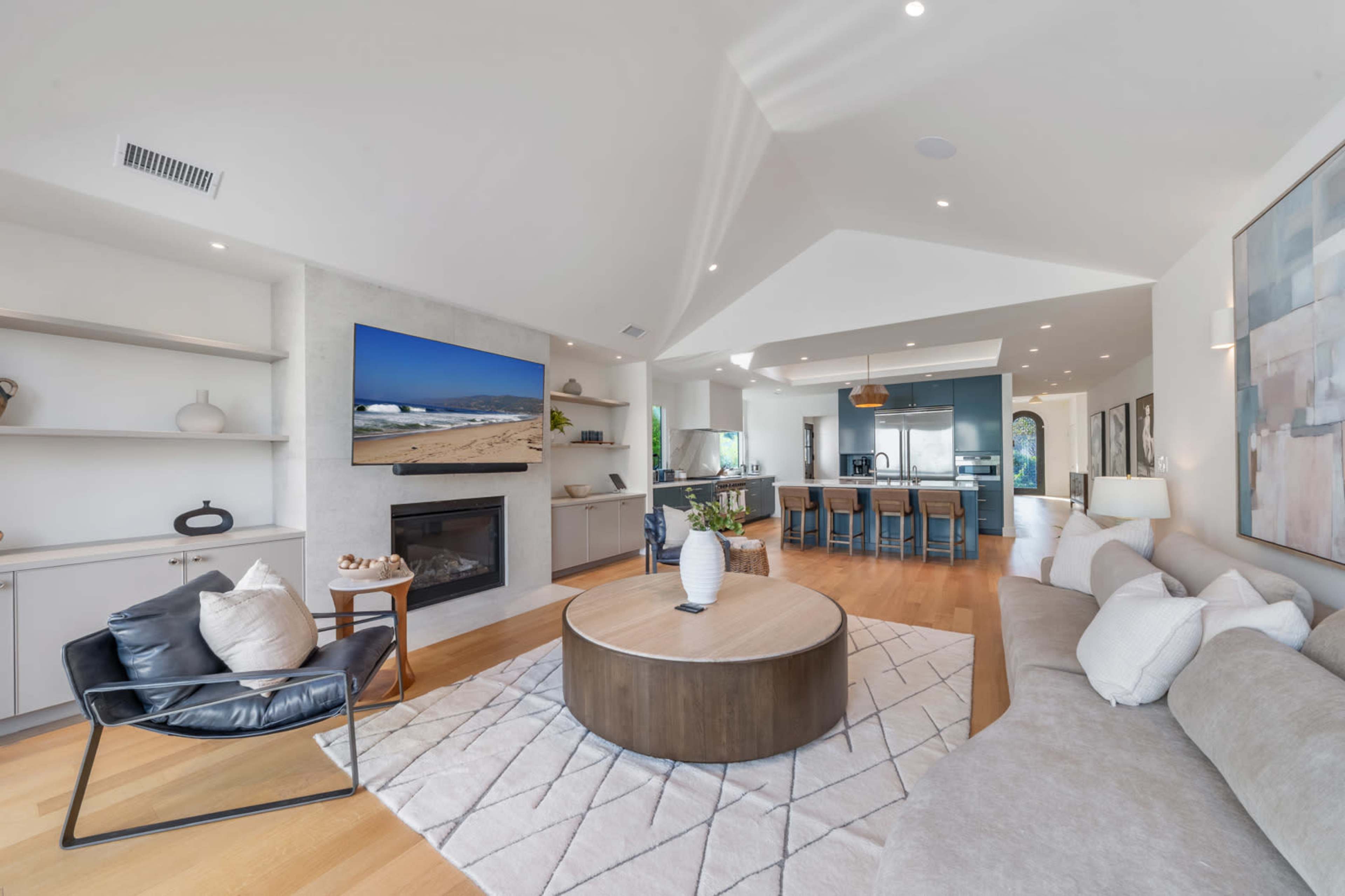 The image shows a modern living room with a large circular coffee table, a sectional sofa, a black lounge chair, a fireplace, and an open kitchen area in the background.