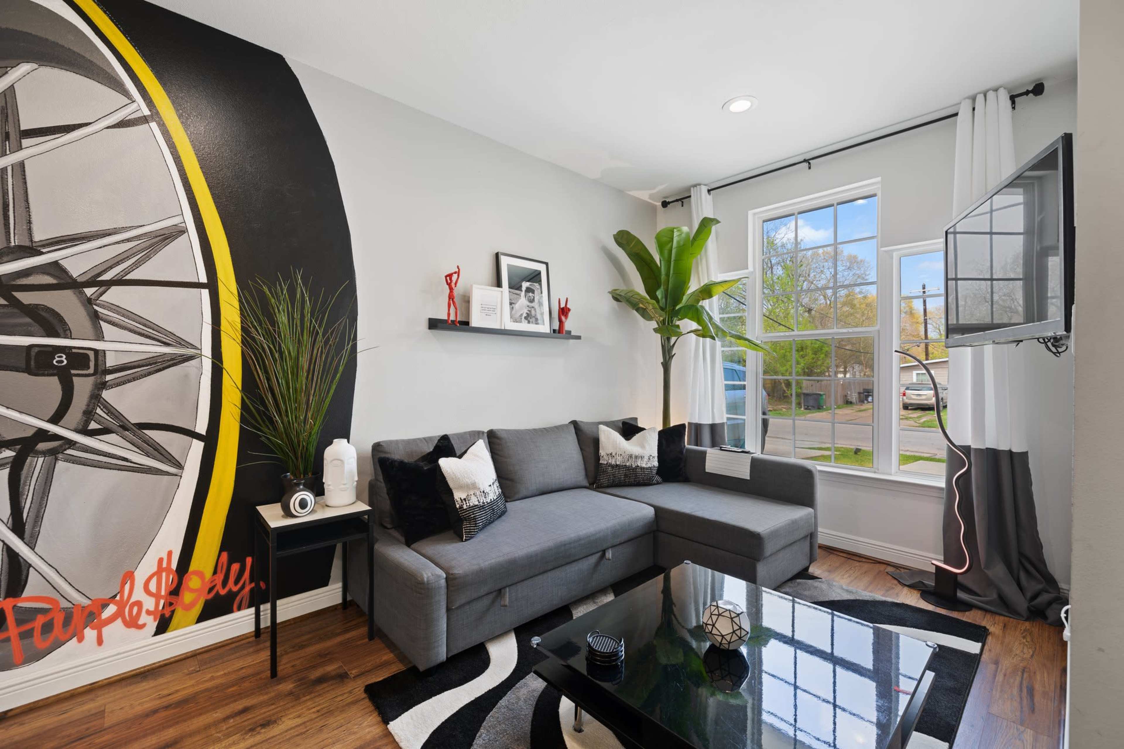 A modern living room features a gray sectional couch, a black accent table, a large wall mural of a wheel, and a television mounted on the wall.