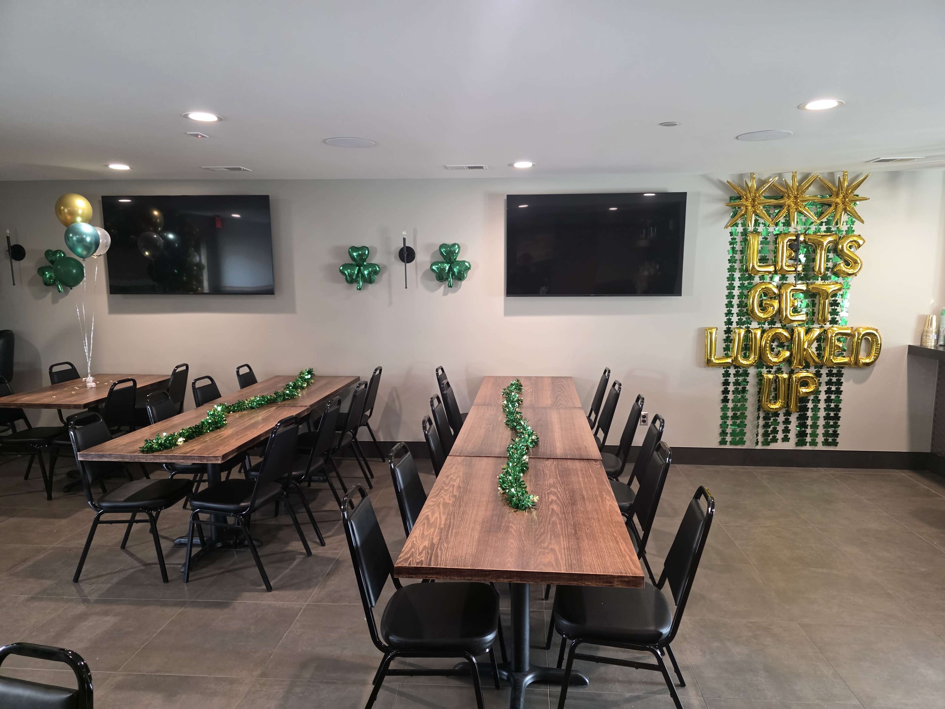 The image shows a decorated event space with two long tables arranged in rows, adorned with green decorations and balloons, while the wall features a "LET'S GET LUCKED UP" display.