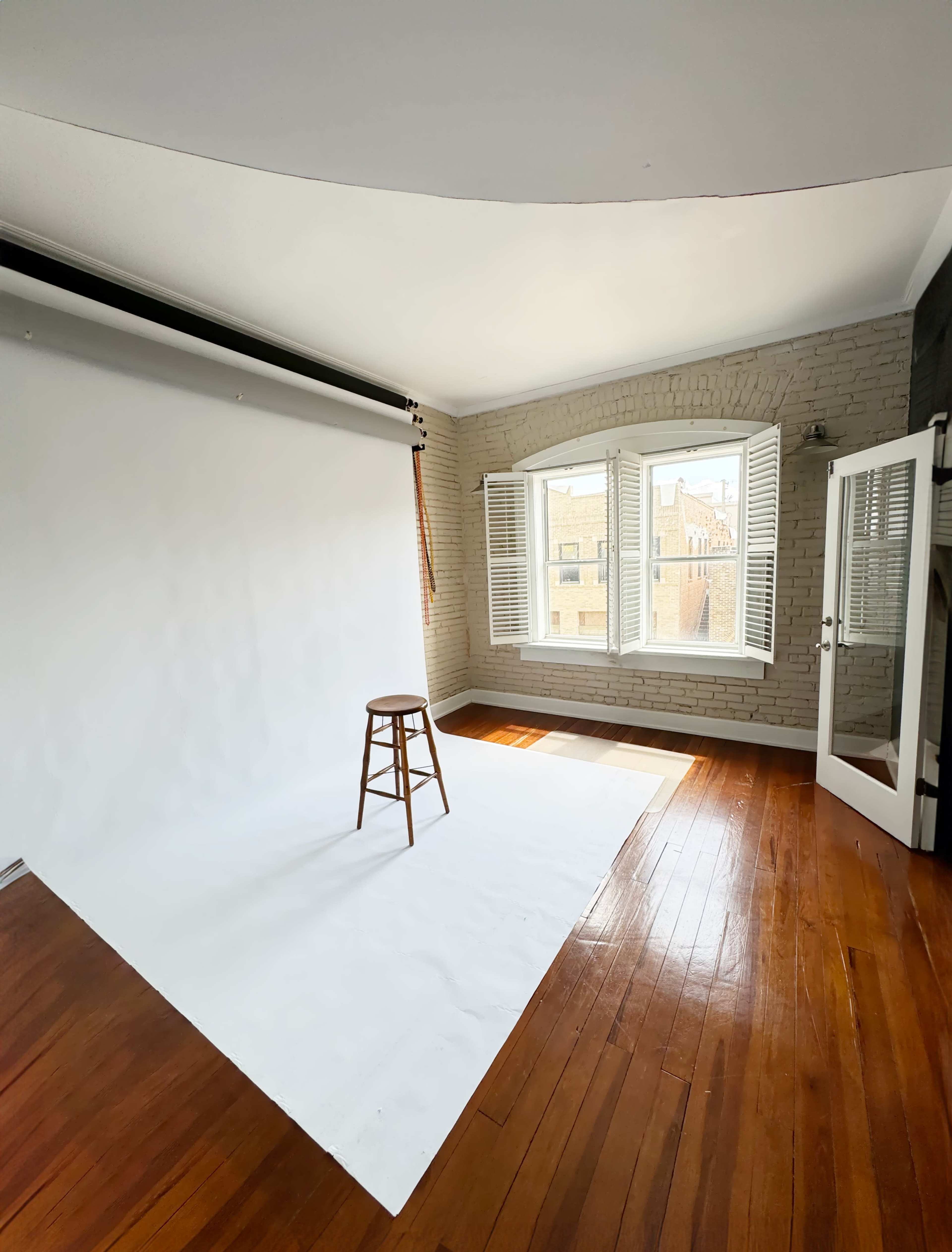 A bright, empty photography studio features a wooden stool on a white backdrop and large windows with shutters.