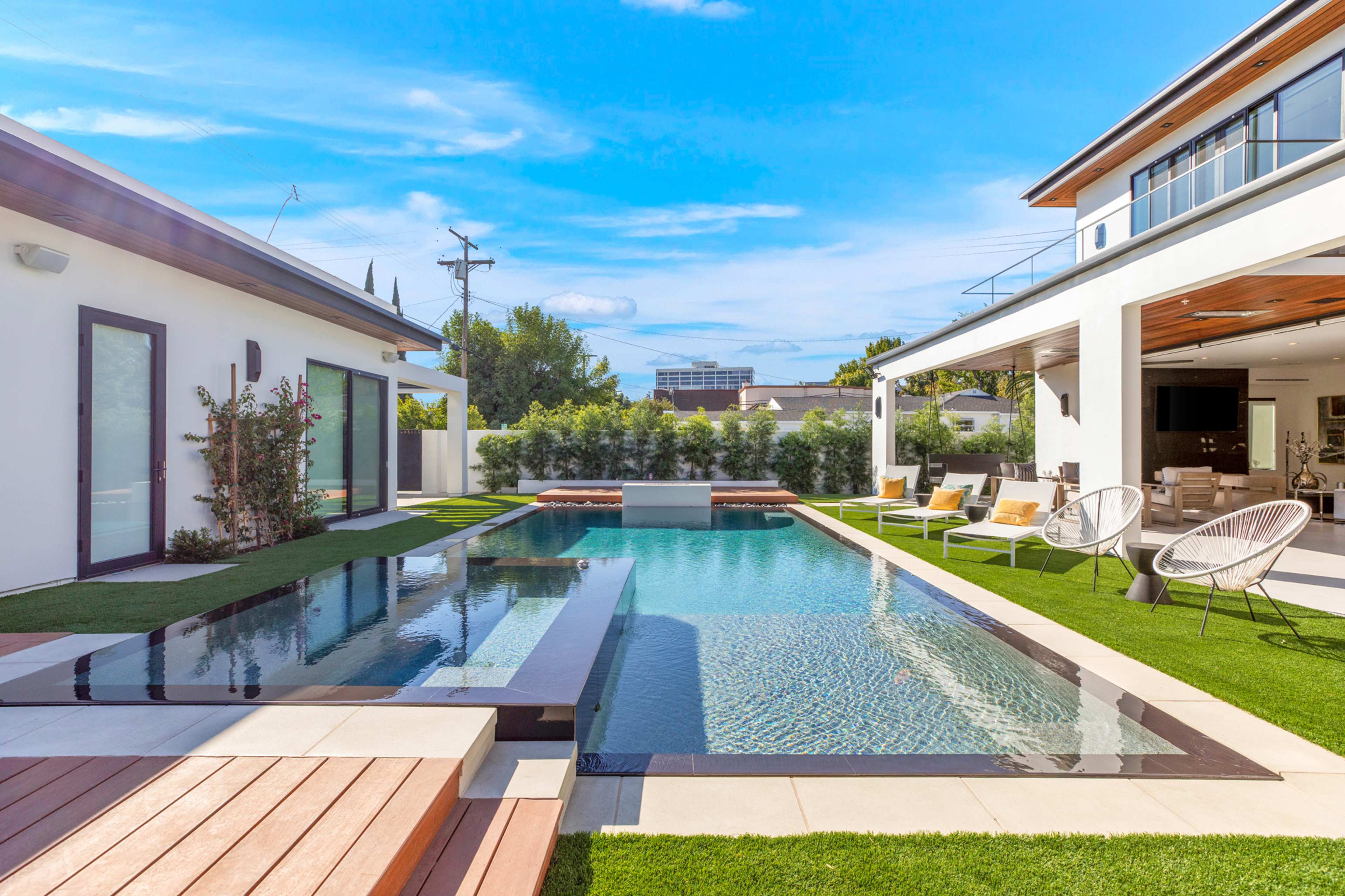 A modern pool area with a rectangular swimming pool surrounded by wooden decking and lounge chairs, adjacent to a stylish house with open outdoor living spaces.