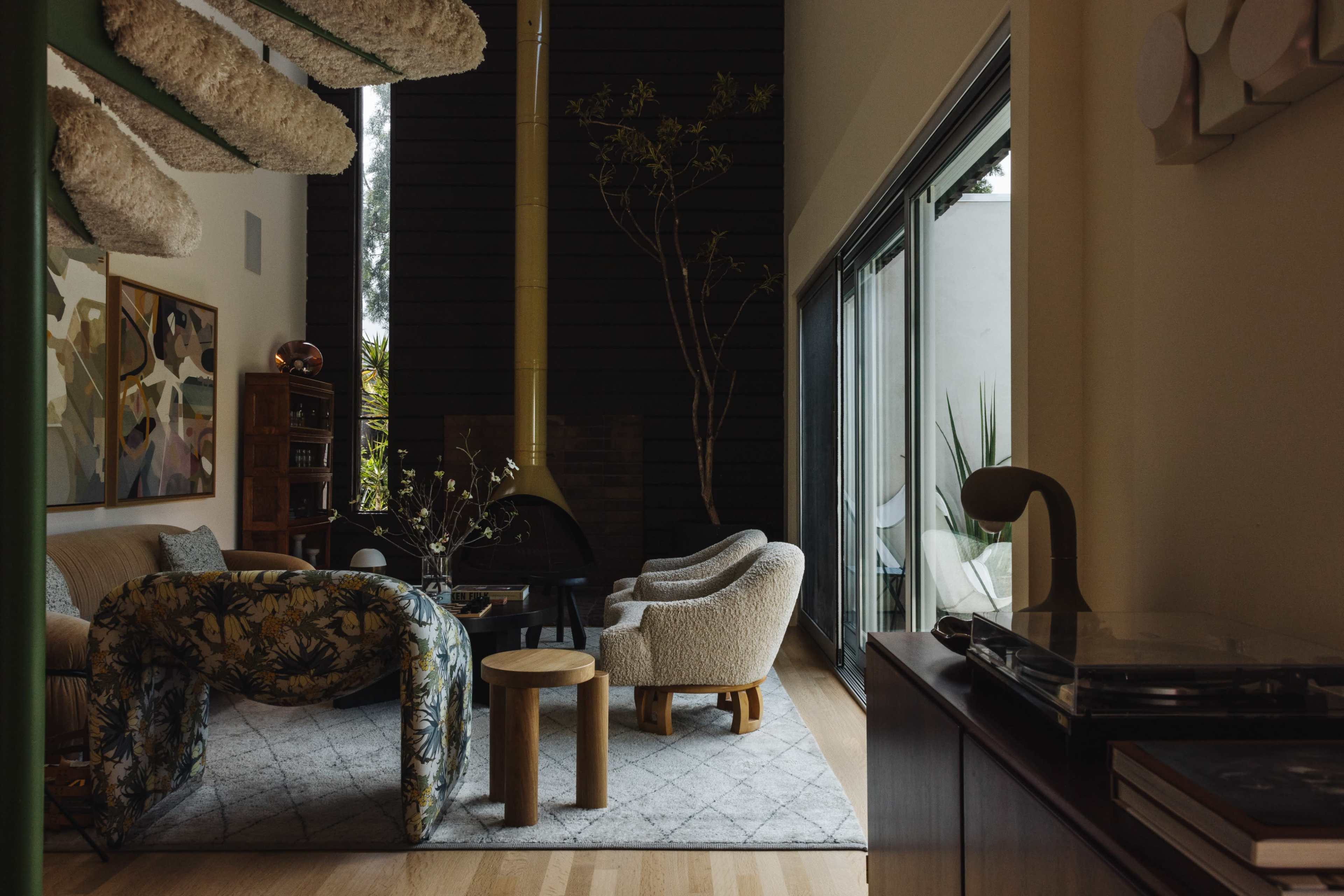 The image shows a modern living room featuring a cozy seating area with a patterned sofa, two armchairs, and a wooden coffee table, alongside large windows that allow natural light to enter.