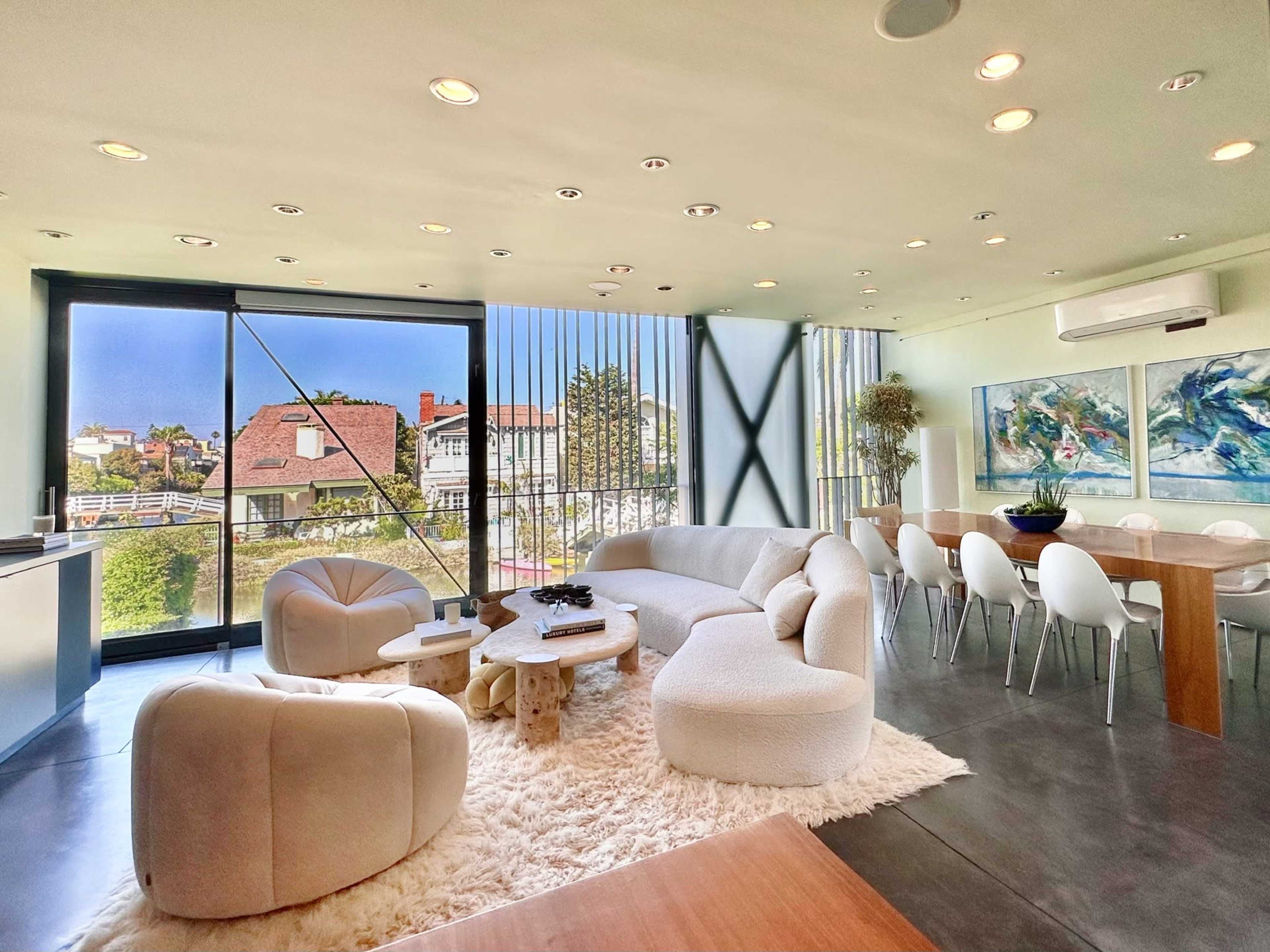 The spacious living area features a cream-colored sofa, a round chair, and a dining table set against large windows overlooking a scenic view.