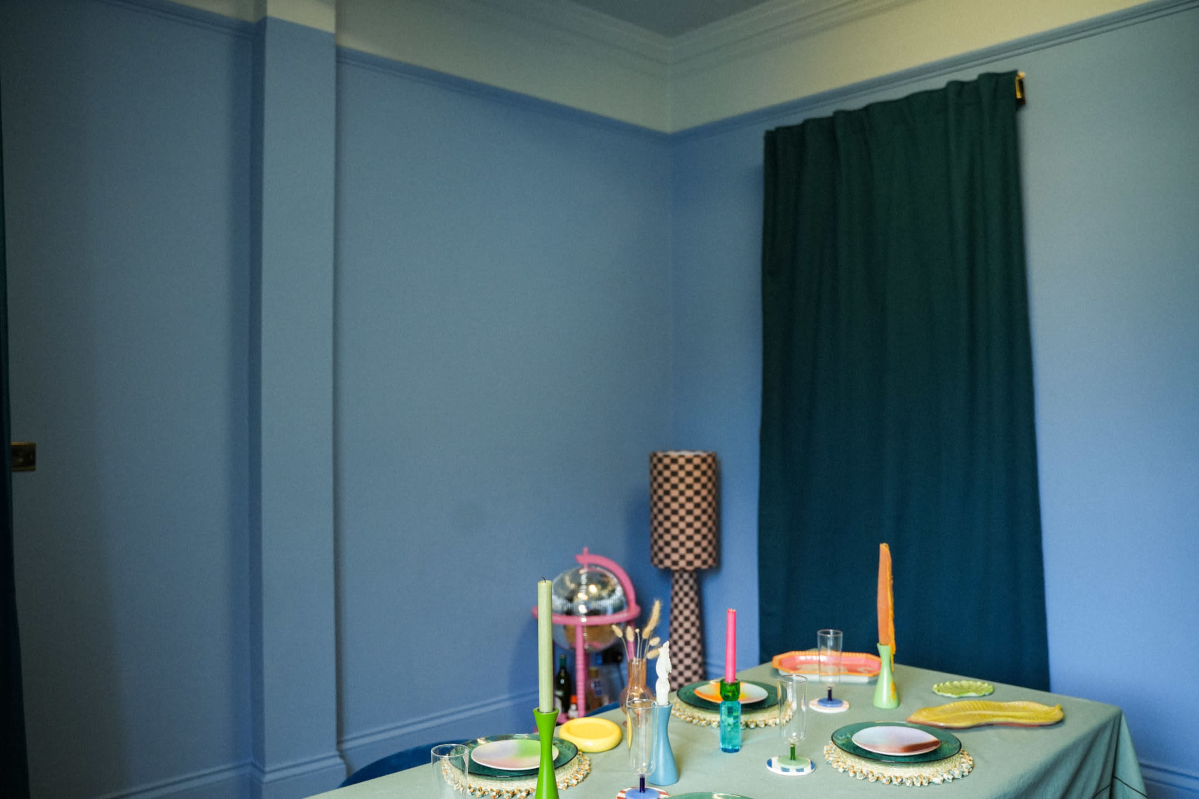 A dining table is set with colorful plates and candles in a room featuring blue walls and a green curtain.