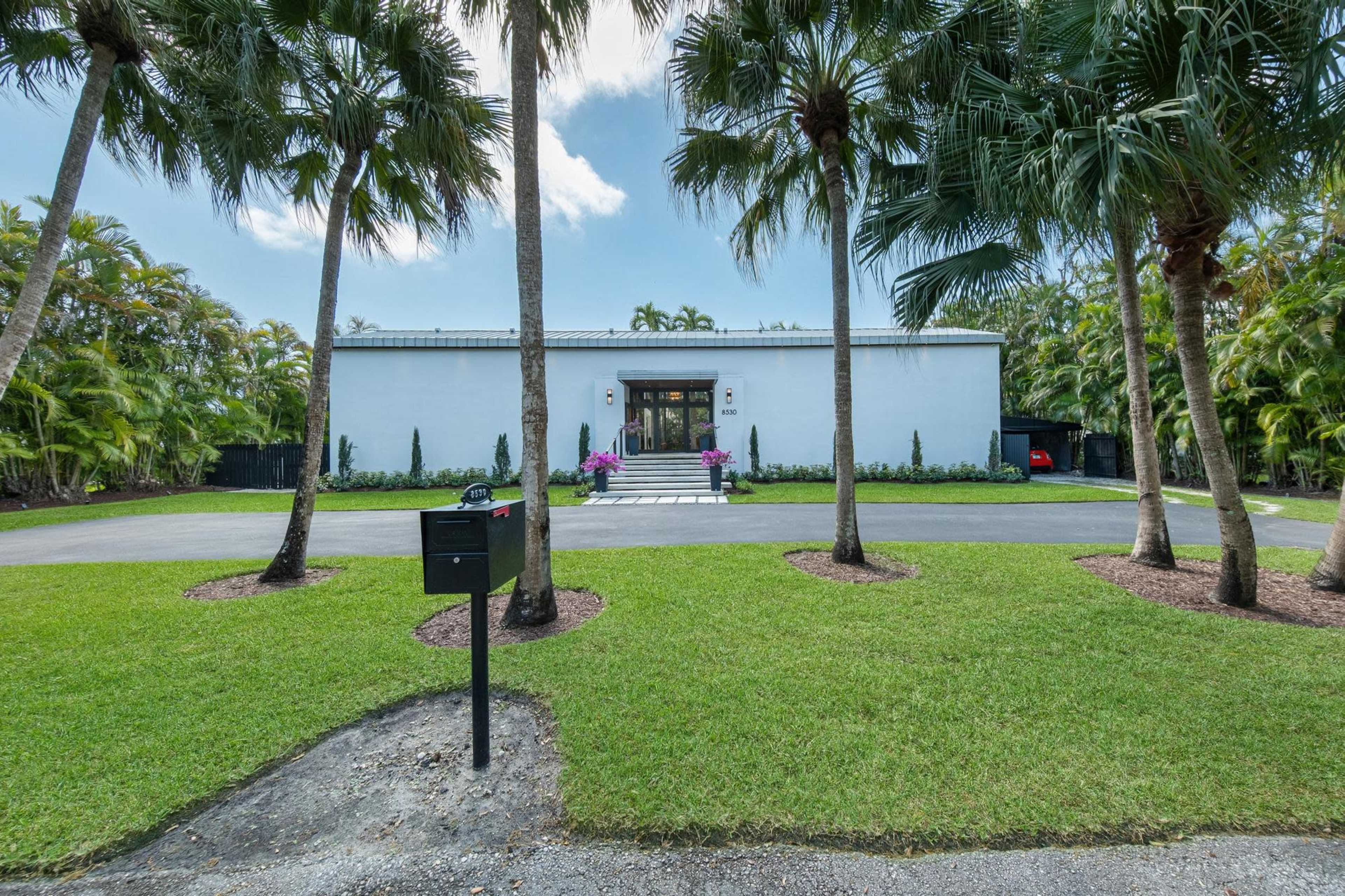 A modern white building with a flat roof is situated behind palm trees, featuring a driveway and landscaped grass in front.