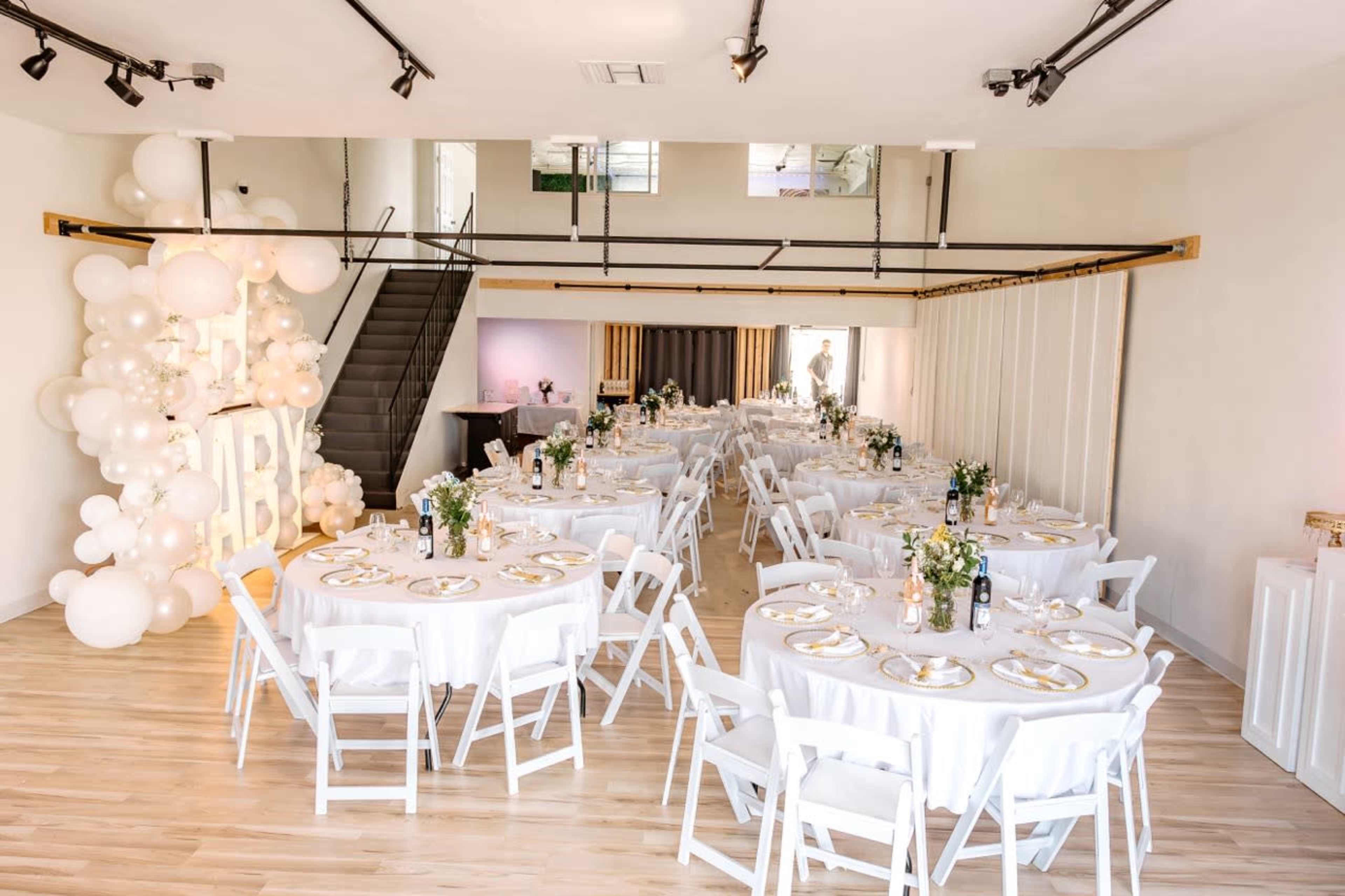 A spacious event venue set with round tables covered in white linens, decorated with floral arrangements, with a backdrop of balloon decor and a staircase in the background.