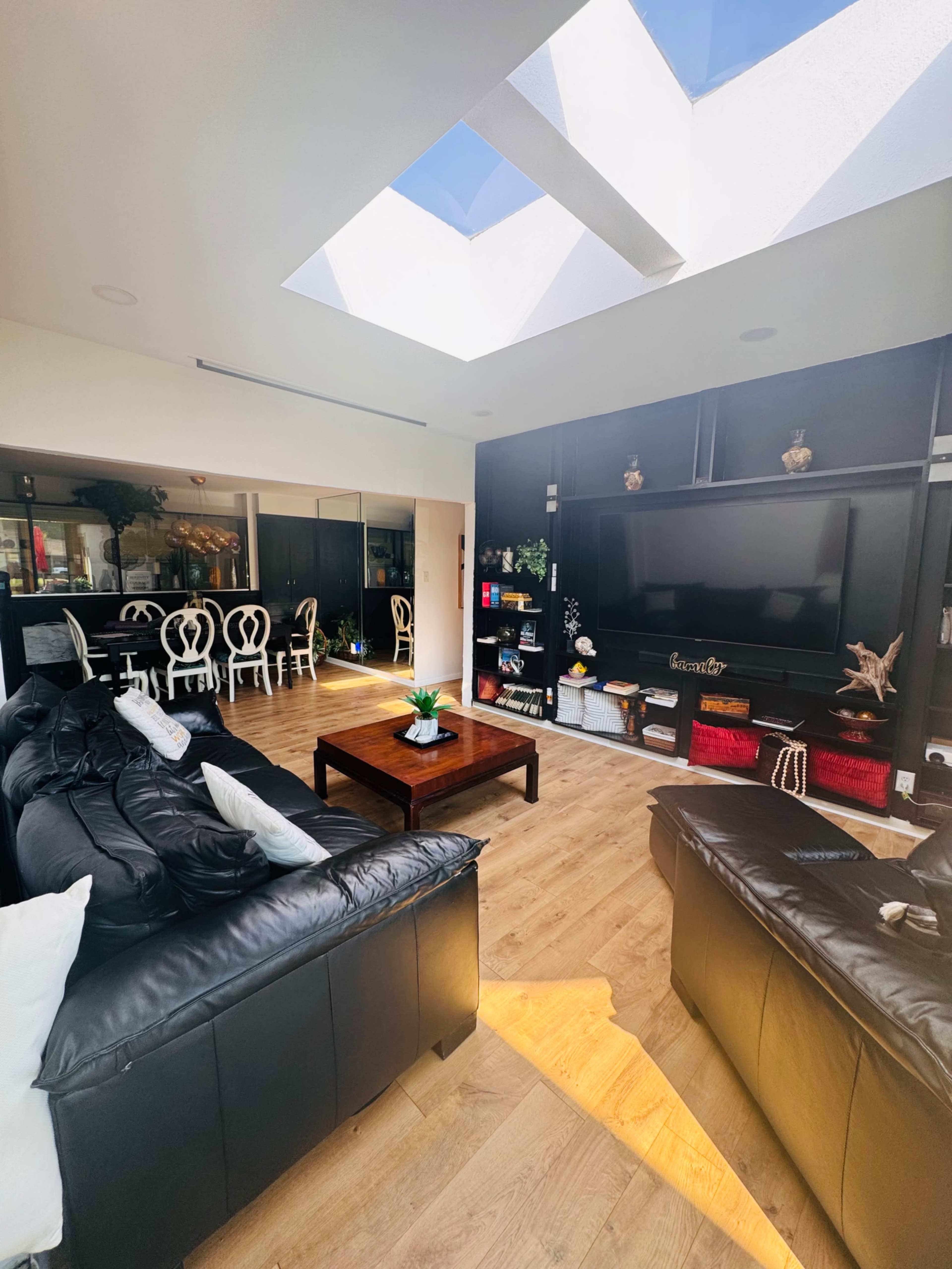 The image shows a bright living room with black leather sofas, a wooden coffee table, and a skylight allowing natural light to fill the space, while a dining area is visible in the background.