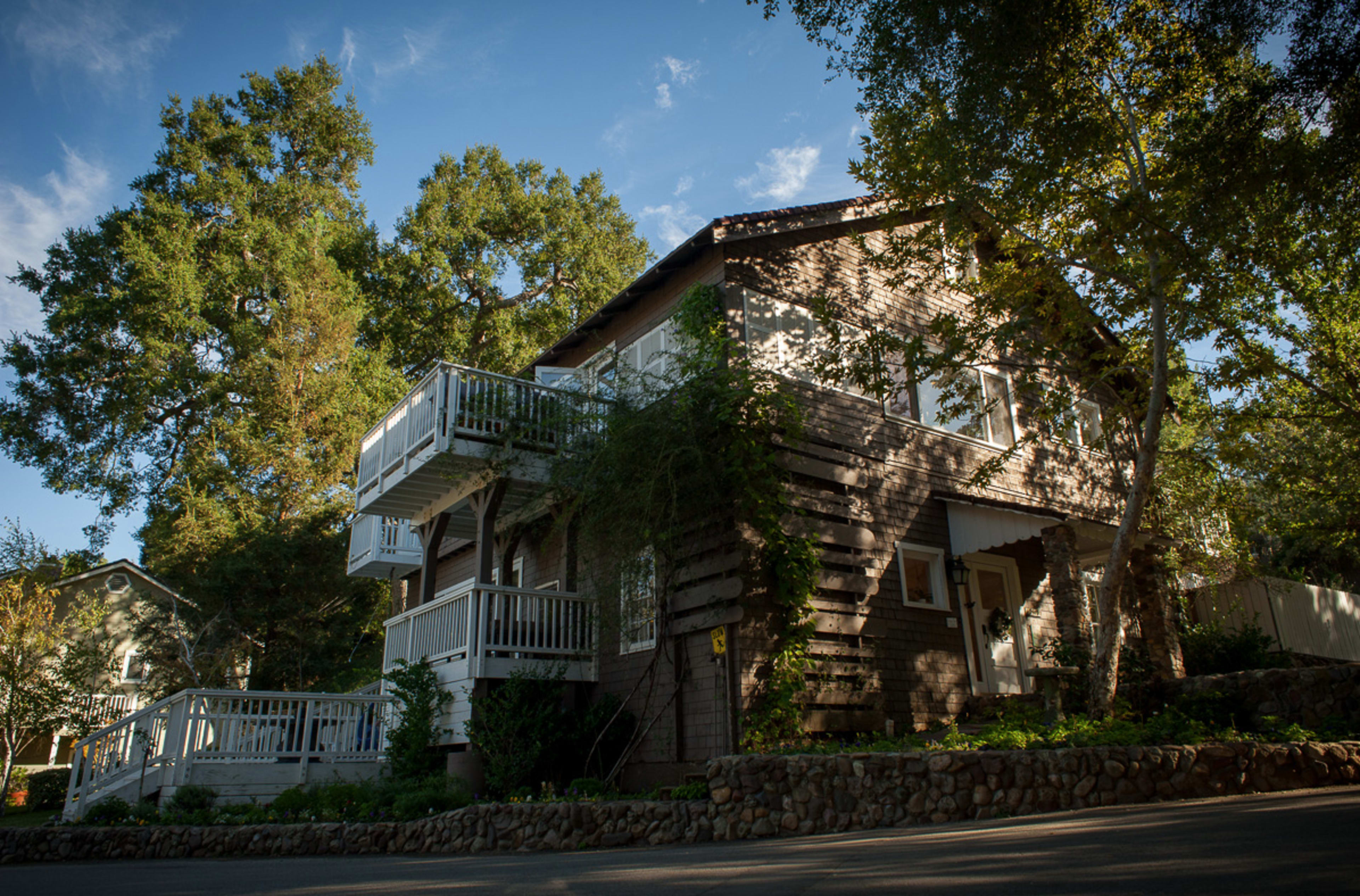 A brown two-story house with a slanted roof and balconies is surrounded by trees and a landscaped front yard.