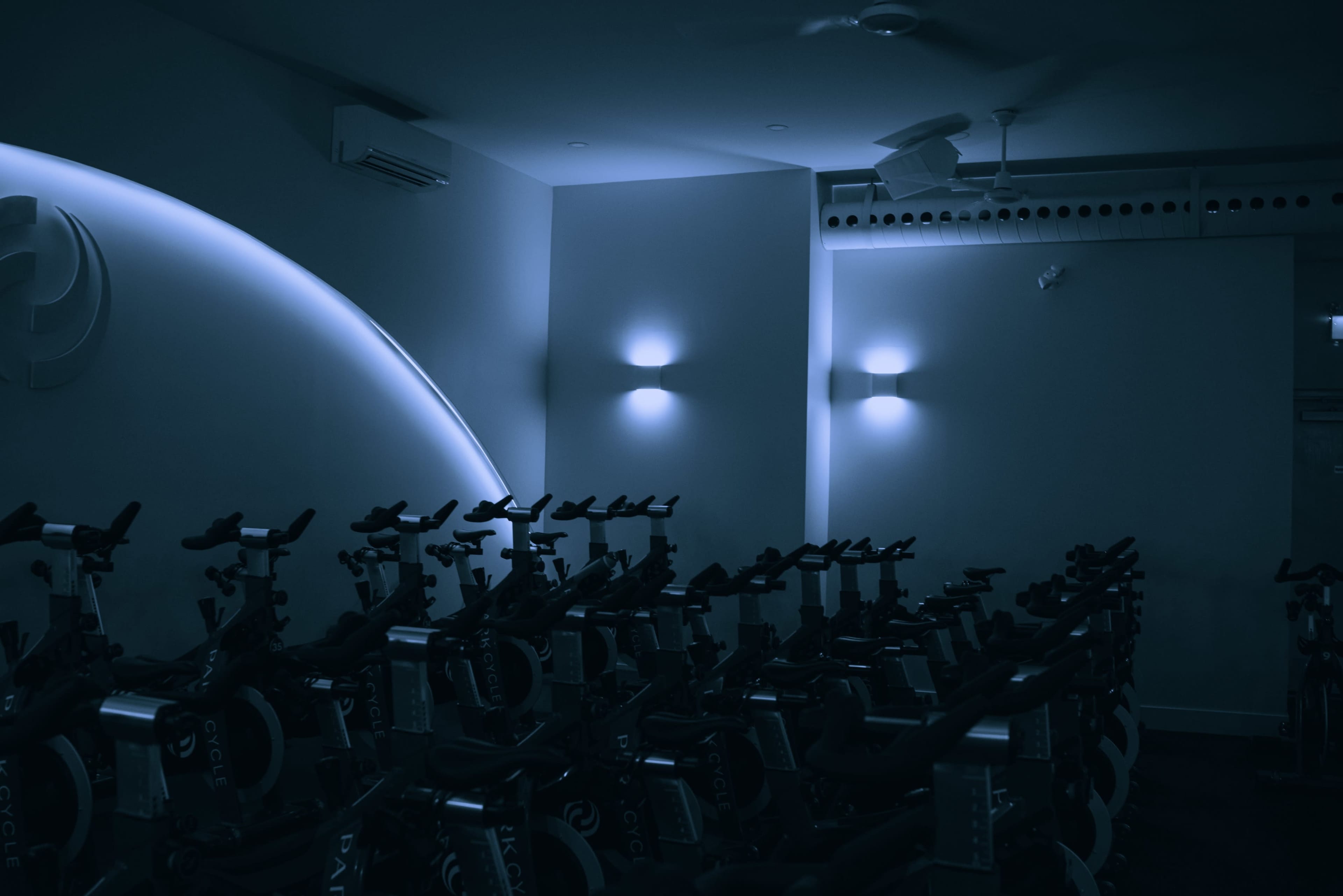 A dimly lit indoor cycling studio features rows of stationary bicycles against a softly illuminated wall.