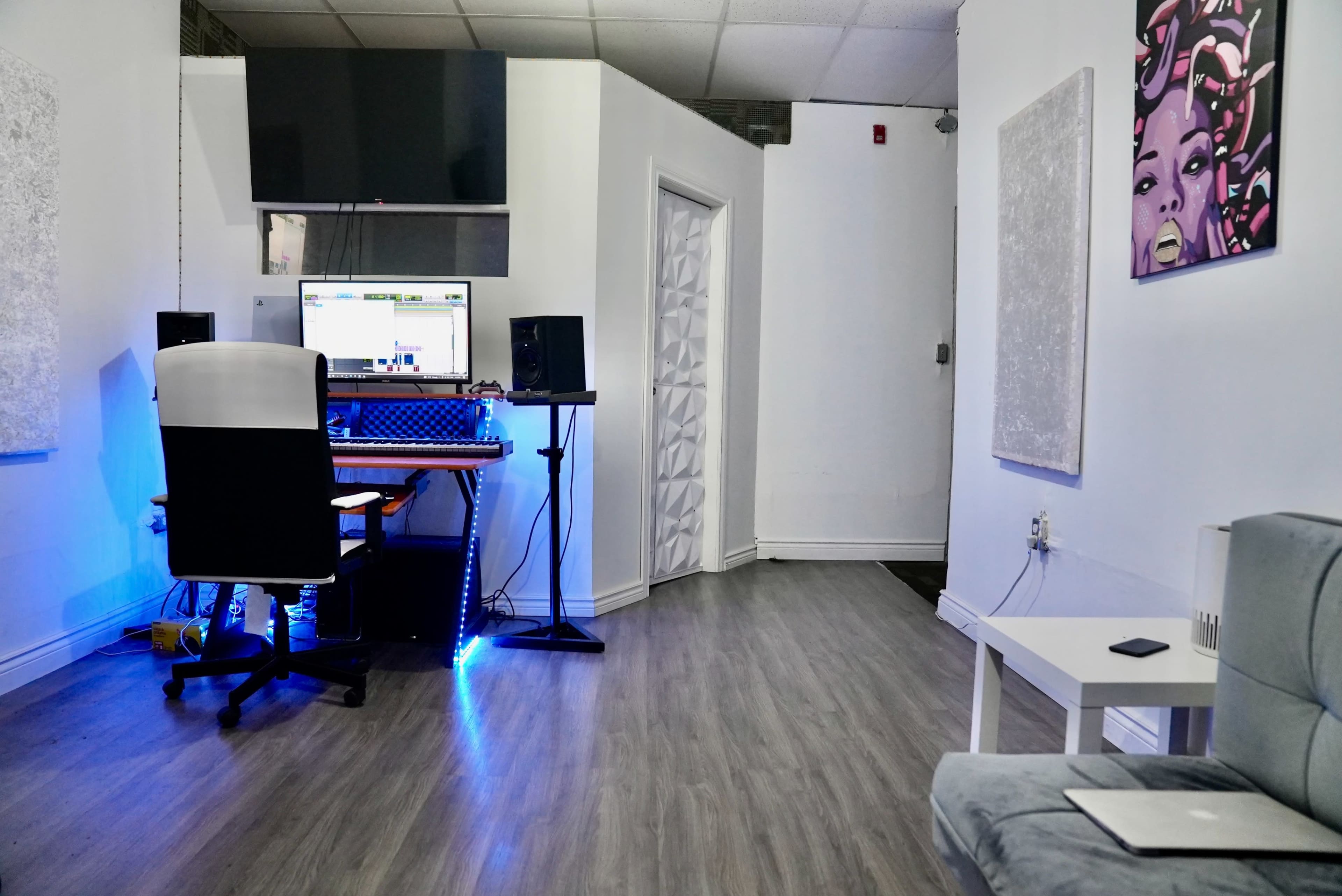 A modern studio space featuring a desk with a computer, audio equipment, and a chair, alongside minimalist decor on the walls and a gray couch in the corner.