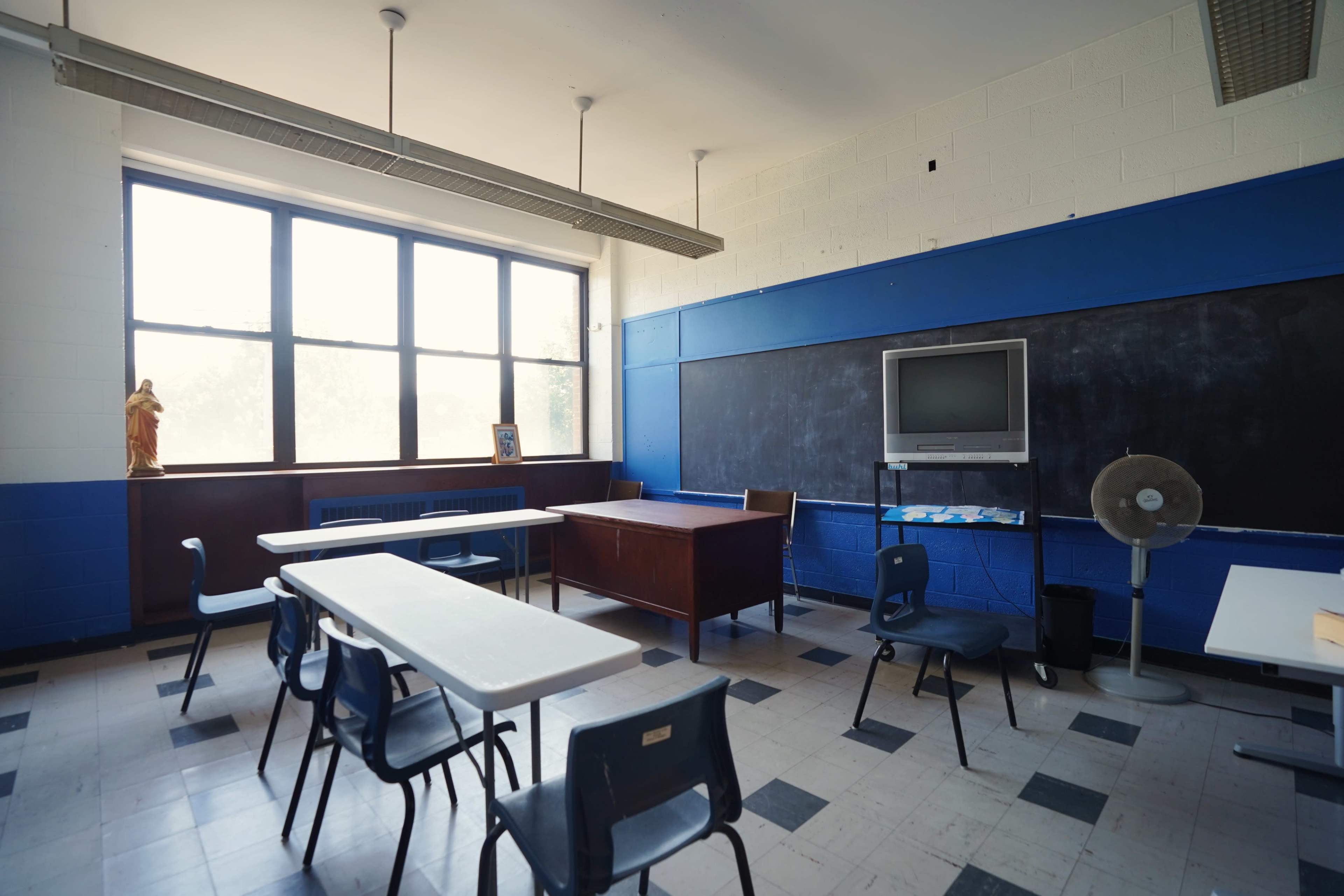 A classroom with blue walls, a wooden desk, several empty chairs, and a chalkboard, illuminated by natural light from large windows.