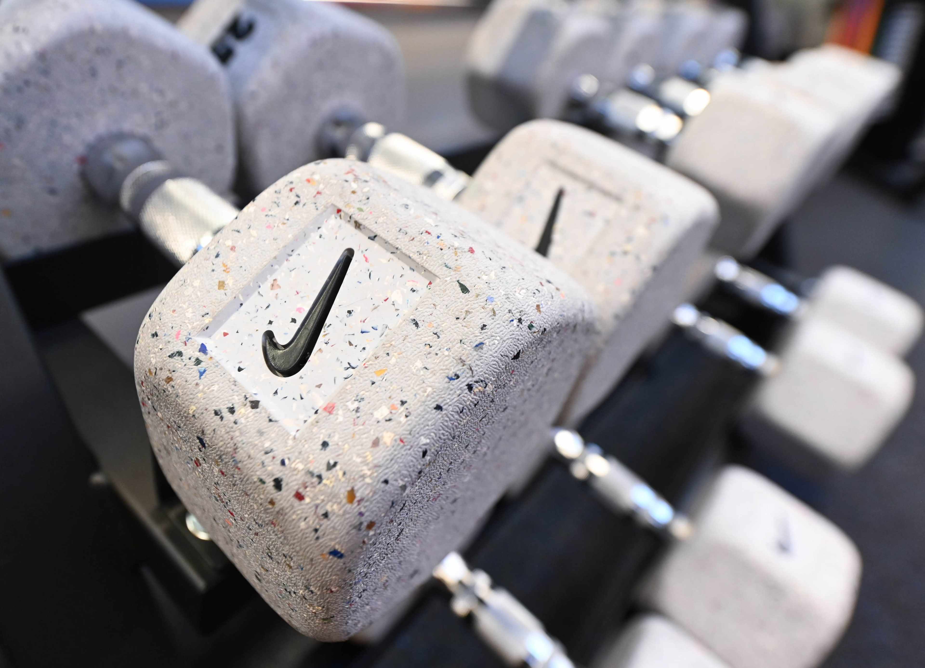 A row of gray dumbbells with the Nike logo sits on a weight rack in a gym.