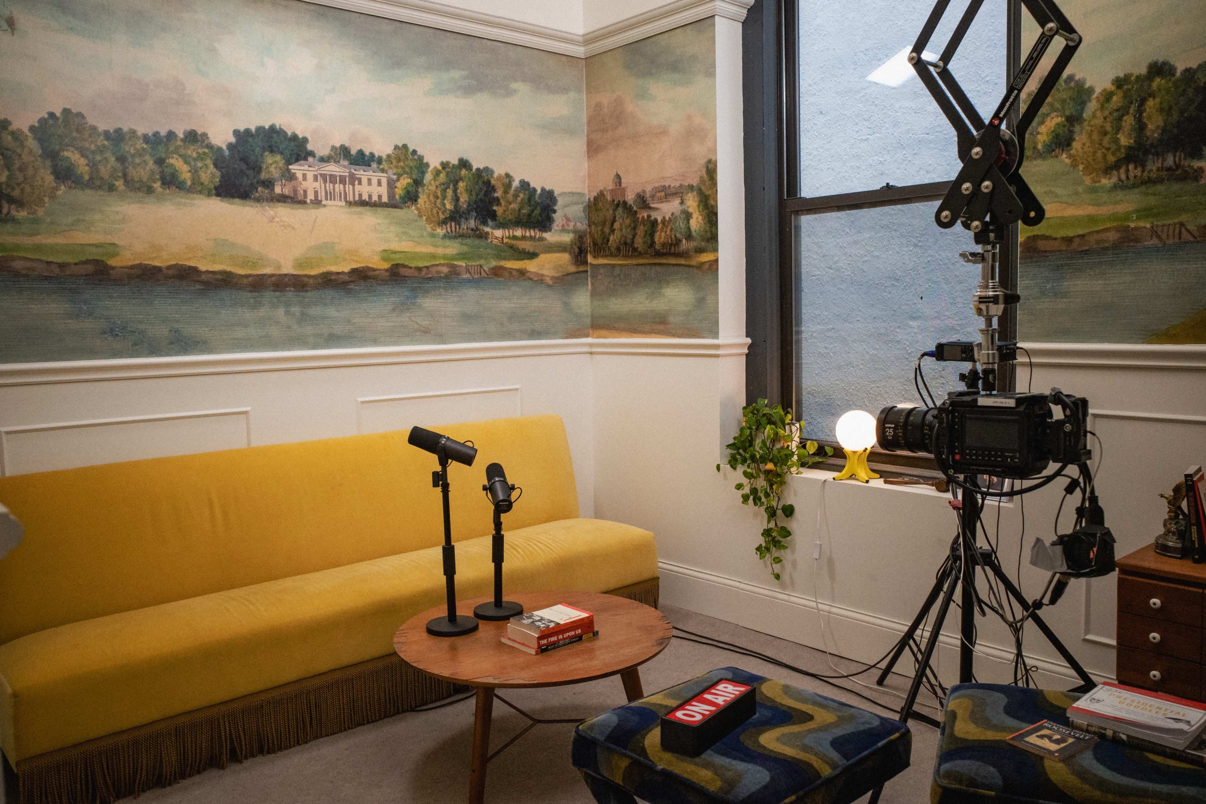 The image shows a cozy room with a yellow sofa, a round coffee table, a microphone set up, and a camera, all surrounded by scenic mural artwork on the walls.