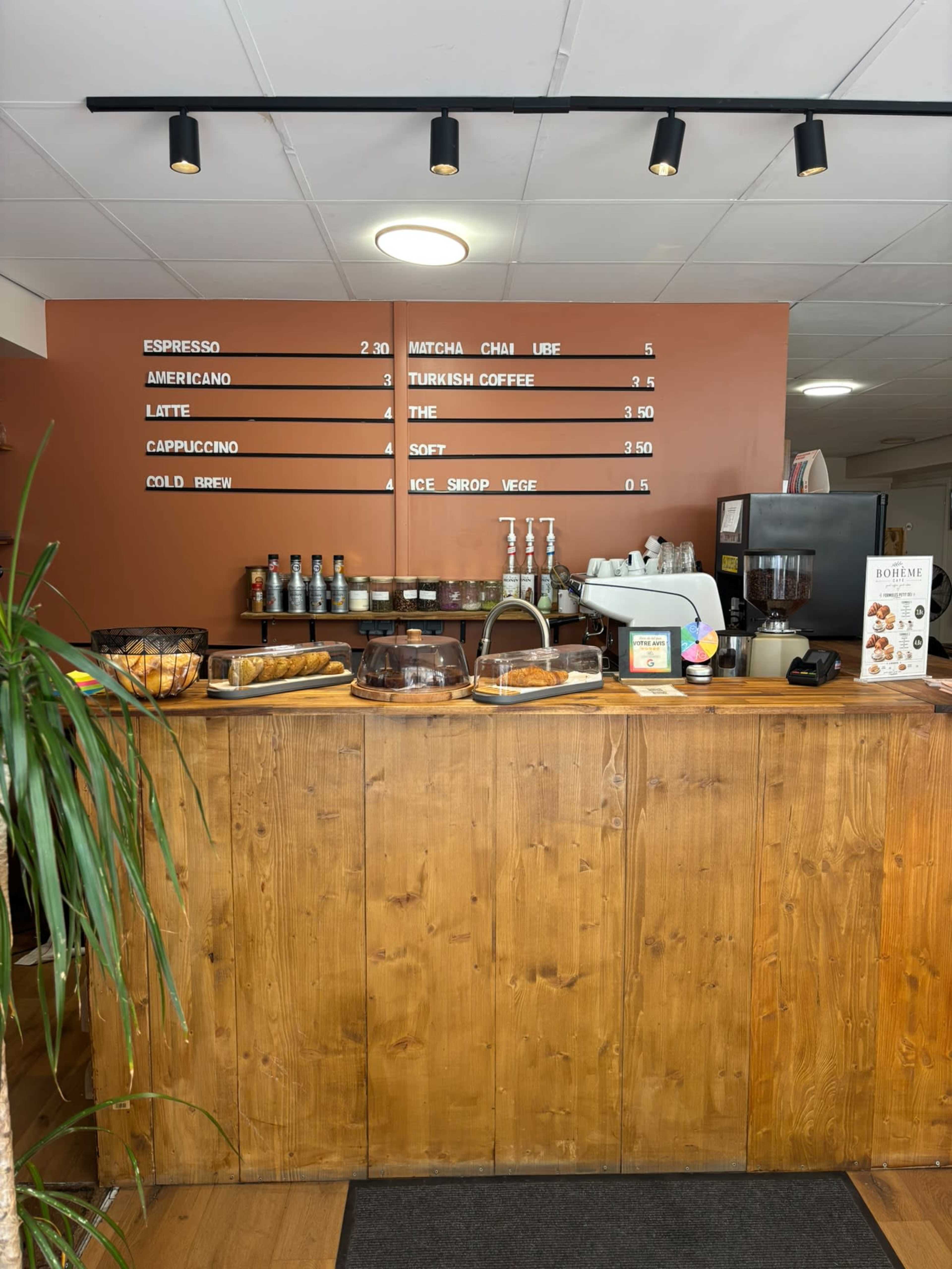 The image shows a coffee shop counter featuring a menu on the wall, a display of baked goods, and various coffee equipment.