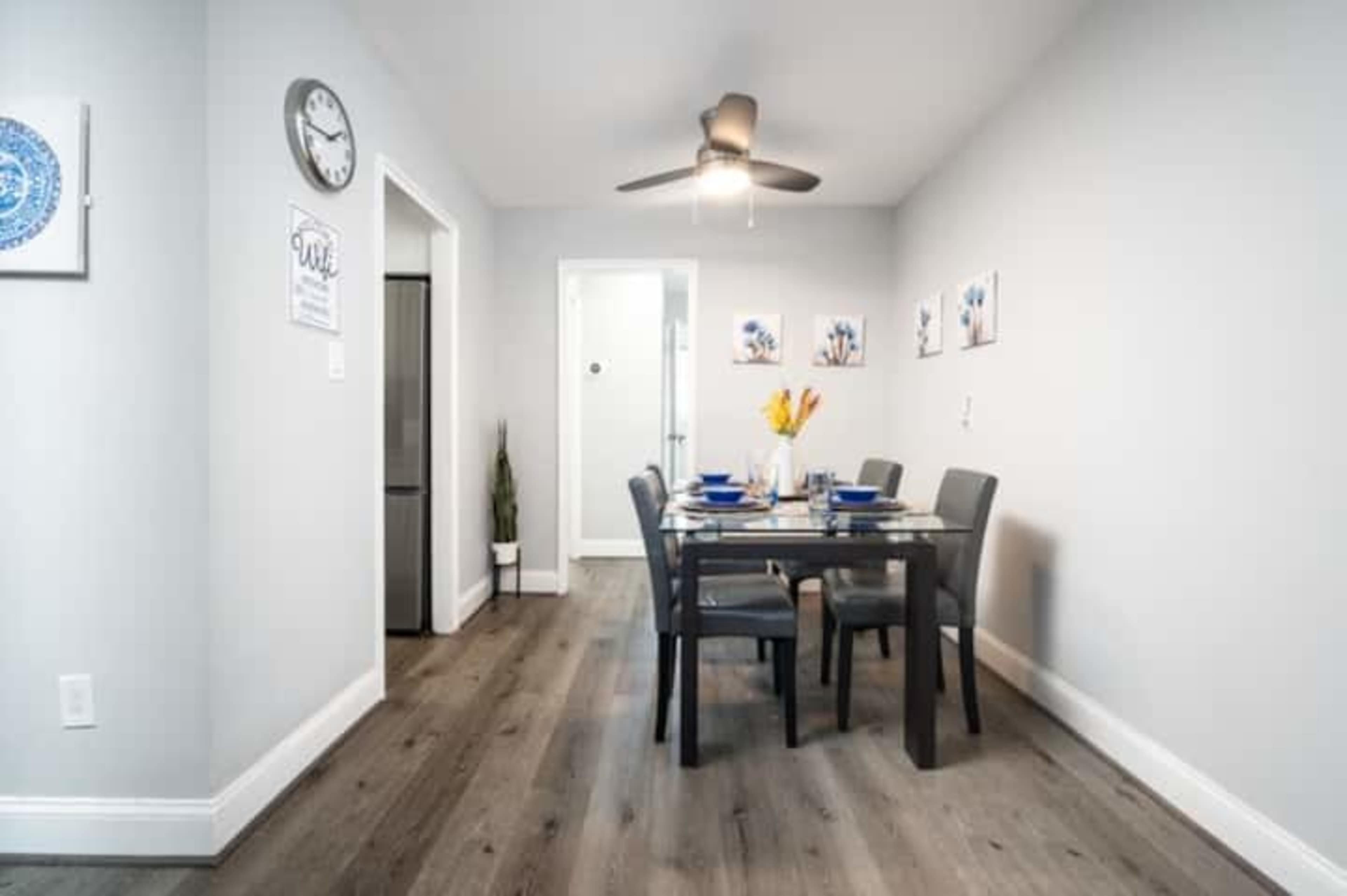 A dining area features a glass table set for four, with a modern light fixture and a wall clock in a well-lit room with gray walls and wooden flooring.