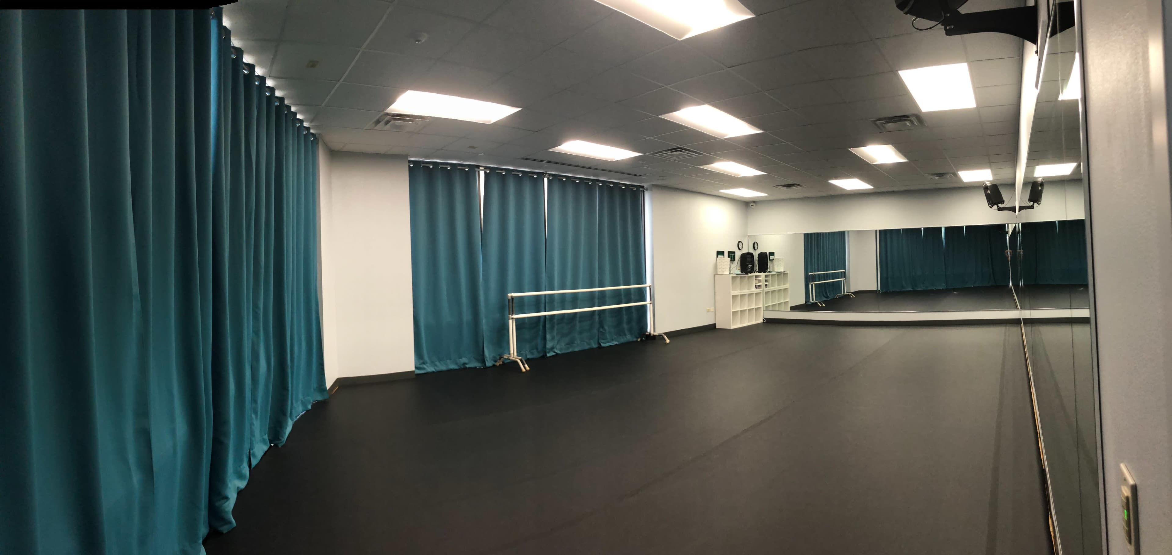 A spacious dance studio with black flooring, teal curtains, and mirrors on one wall.