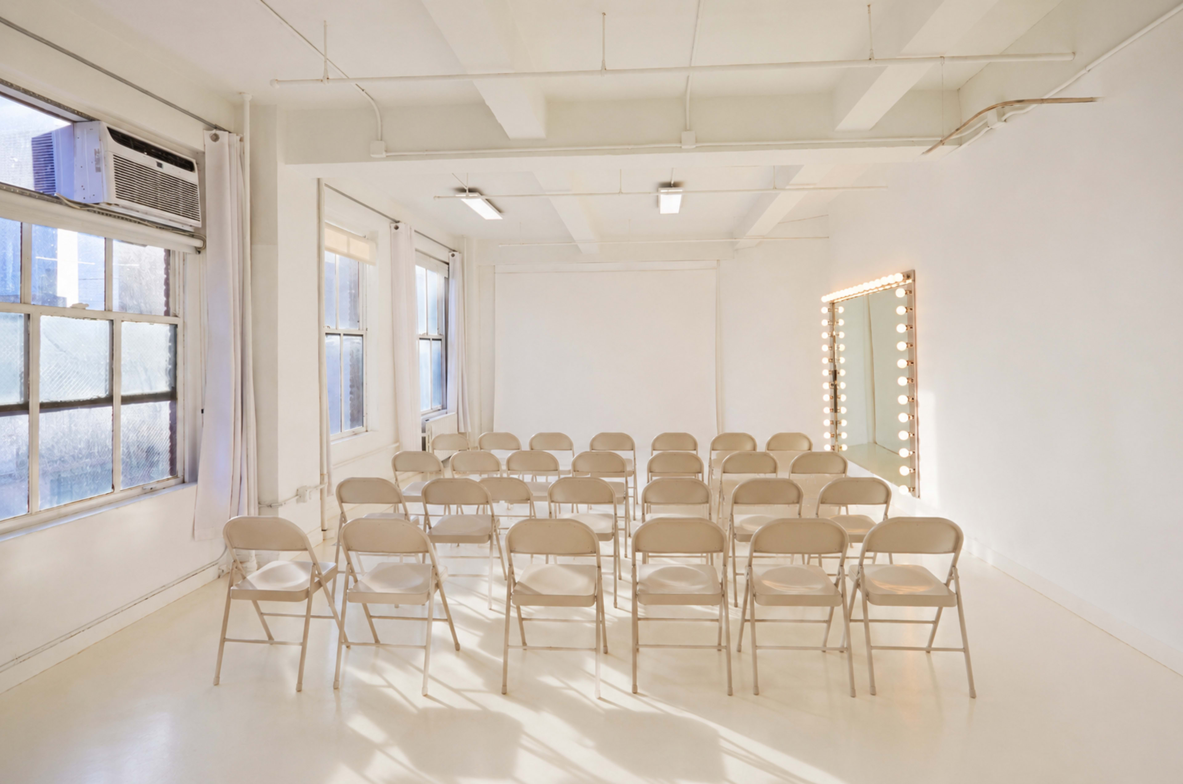 The image shows a bright, empty room with beige folding chairs arranged in rows and a illuminated vanity mirror on one wall.