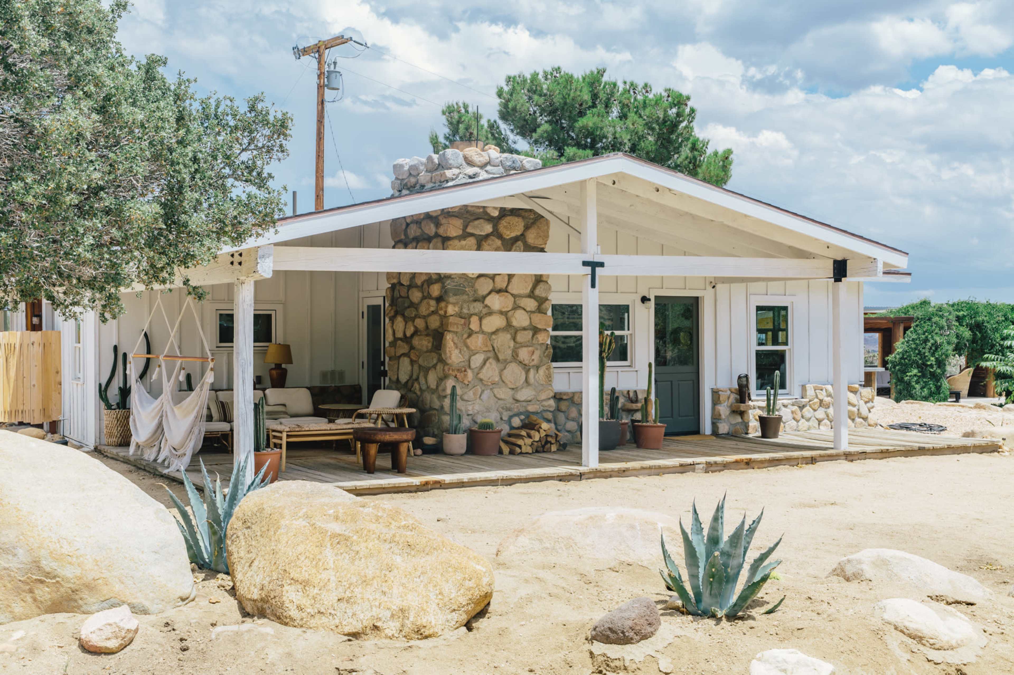 A modern country house with a stone chimney, surrounded by large rocks and desert vegetation, features a covered porch and outdoor seating.