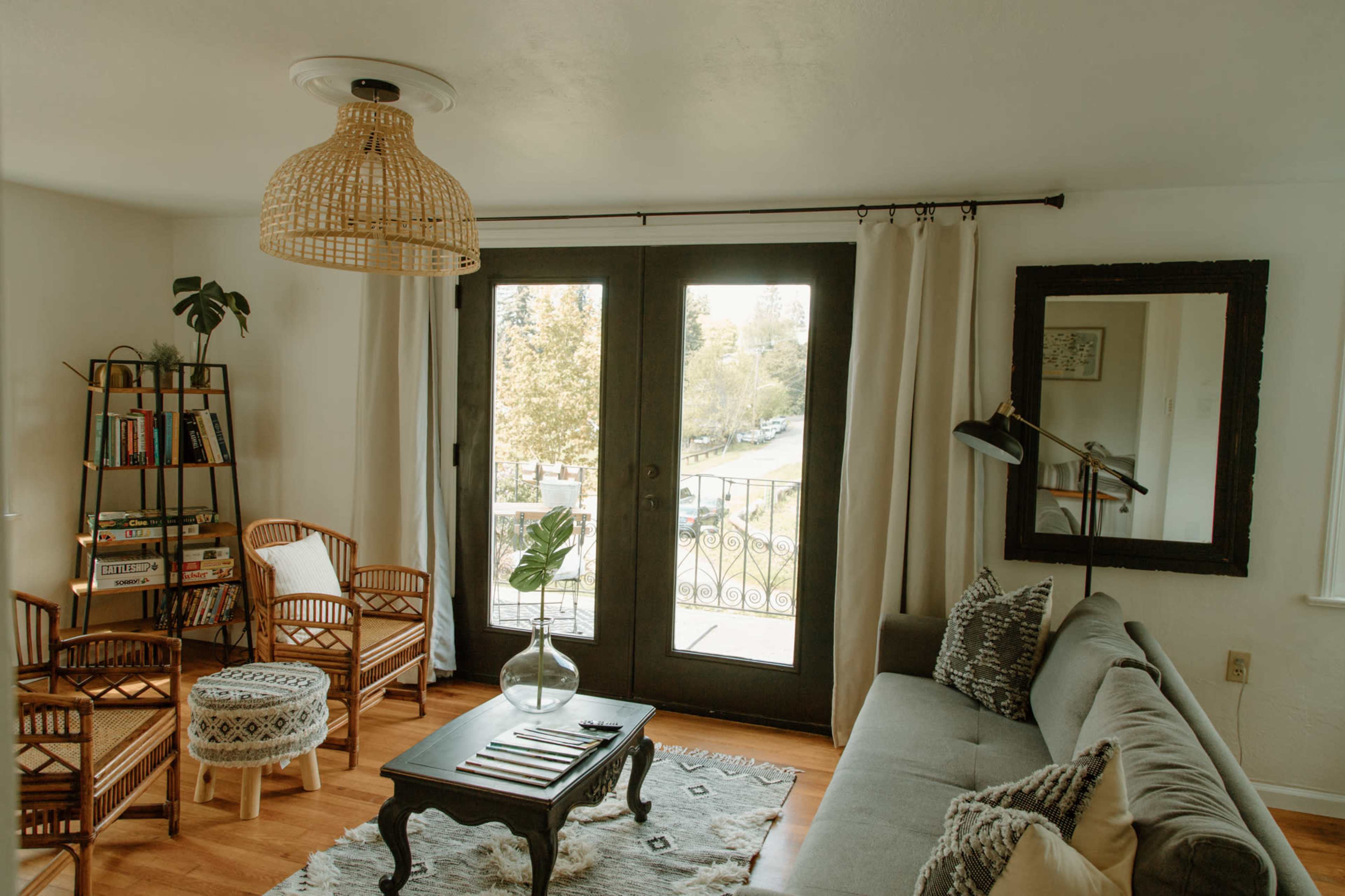 The image shows a cozy living room with a gray sofa, rattan chairs, a wooden coffee table, and large doors that open to a balcony overlooking a tree-lined view.