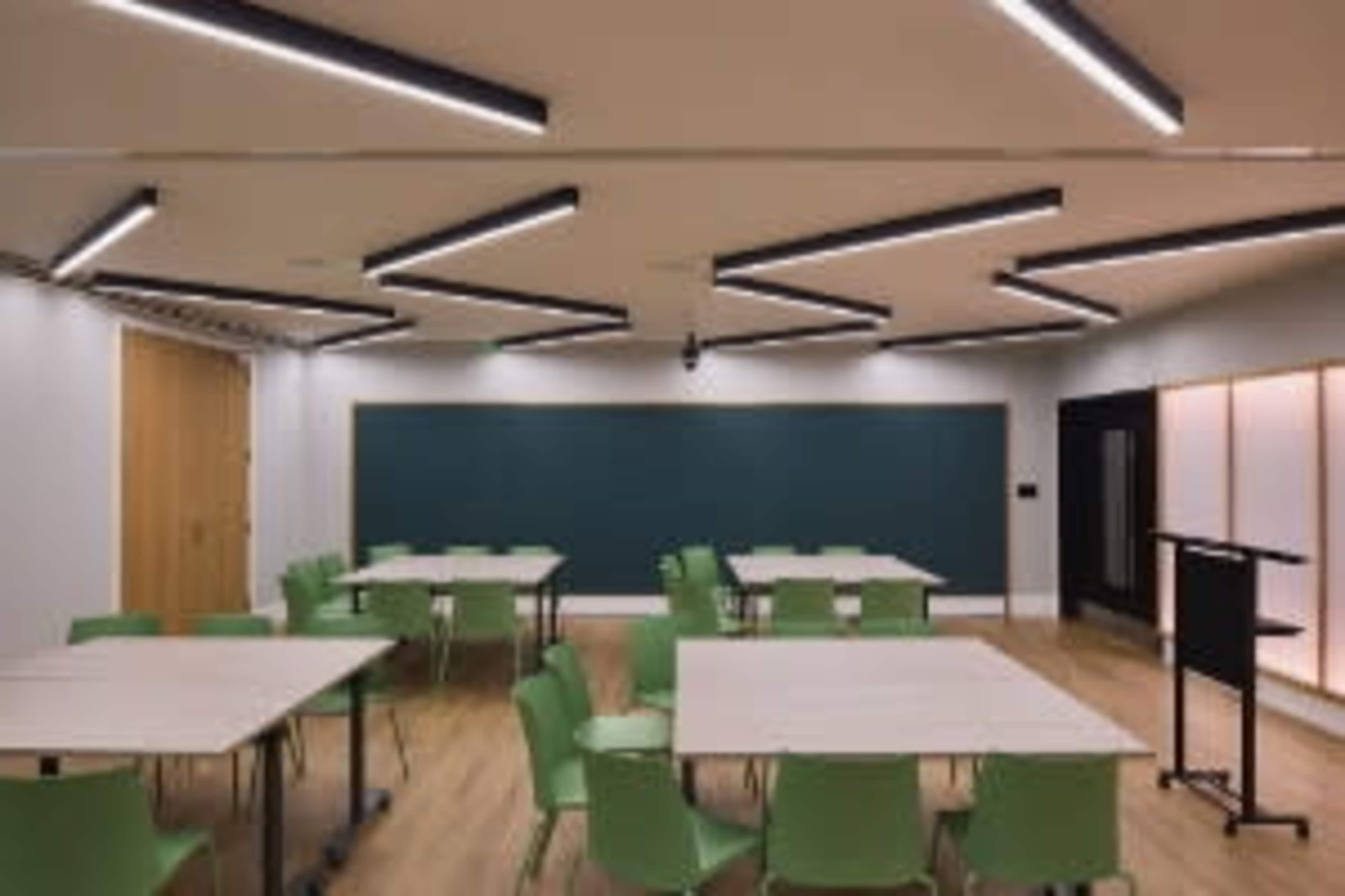 The image shows a modern classroom with green chairs and wooden tables arranged in a grid, a chalkboard on one wall, and unique, angular lighting fixtures on the ceiling.
