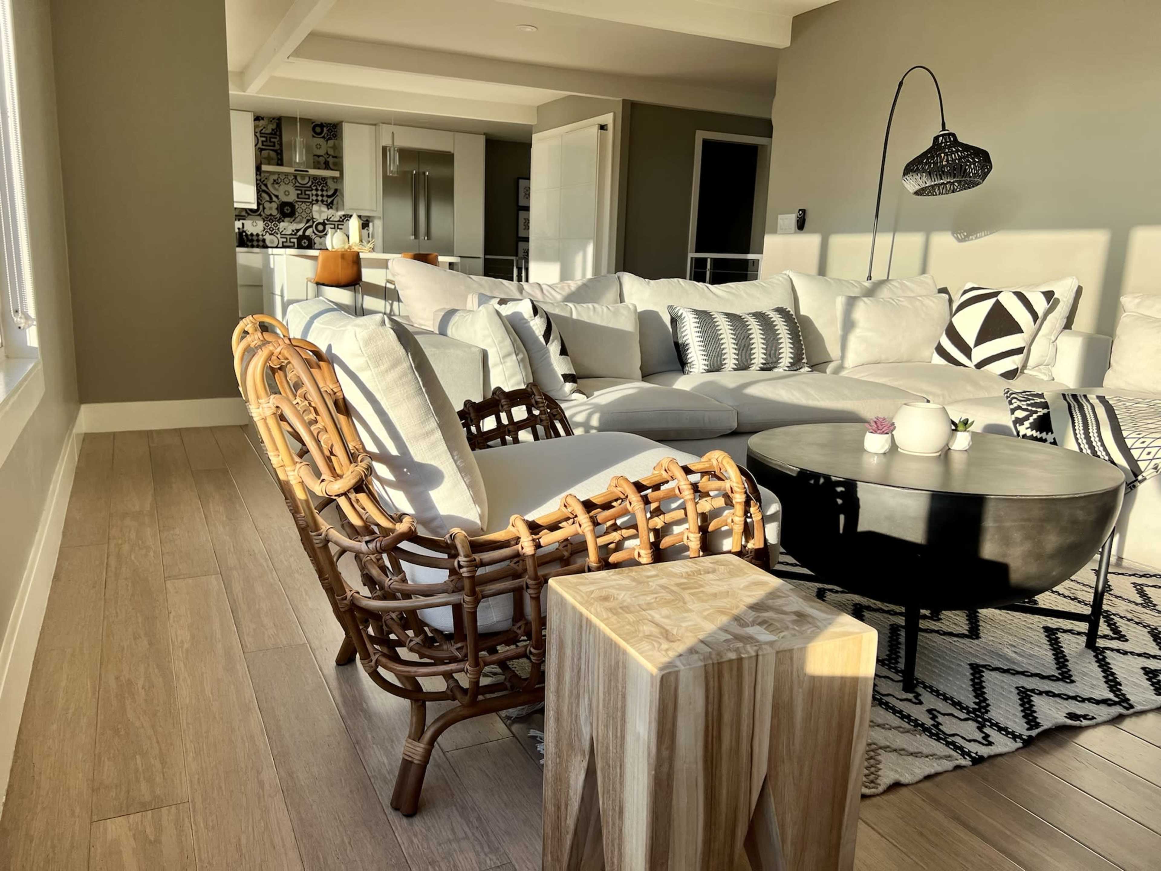 The image shows a modern living room with a sectional sofa, a round coffee table, a rattan chair, and a wooden stool, all arranged on hardwood flooring.