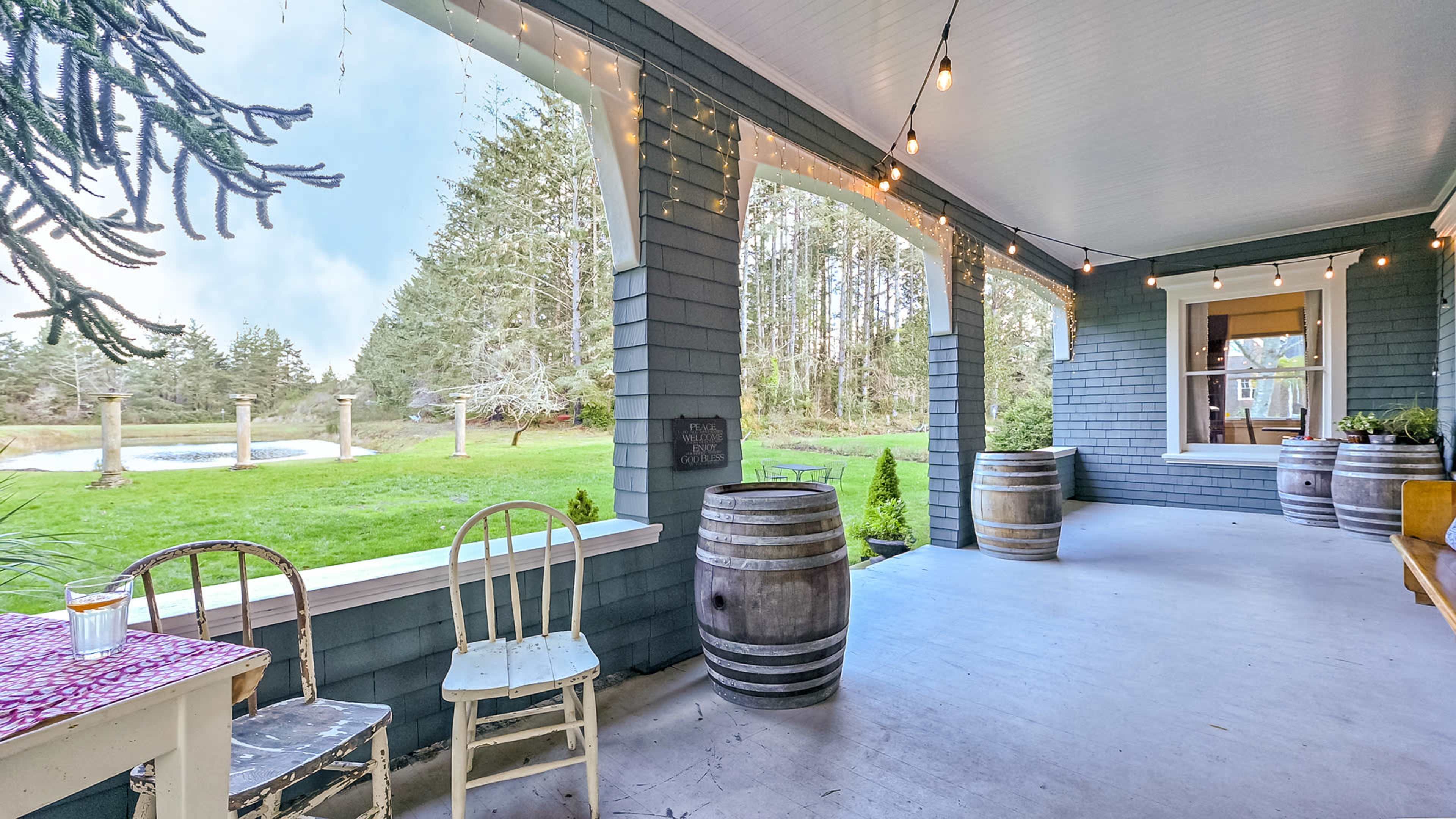 A porch with two wooden chairs and wine barrels, decorated with string lights, overlooking a green lawn and trees in the background.