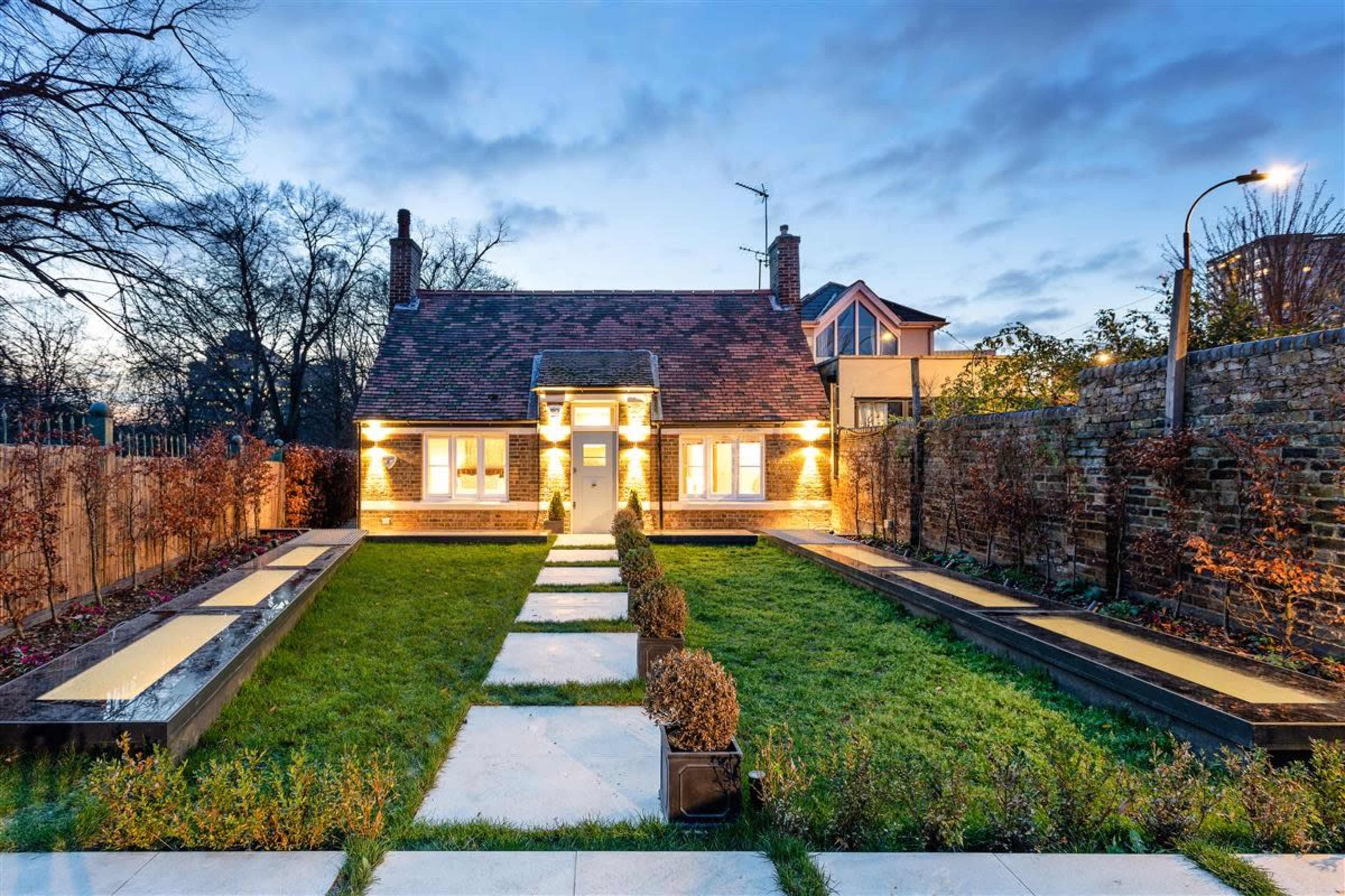 A well-lit house with a manicured lawn and walkways, surrounded by landscaped gardens and a stone wall.