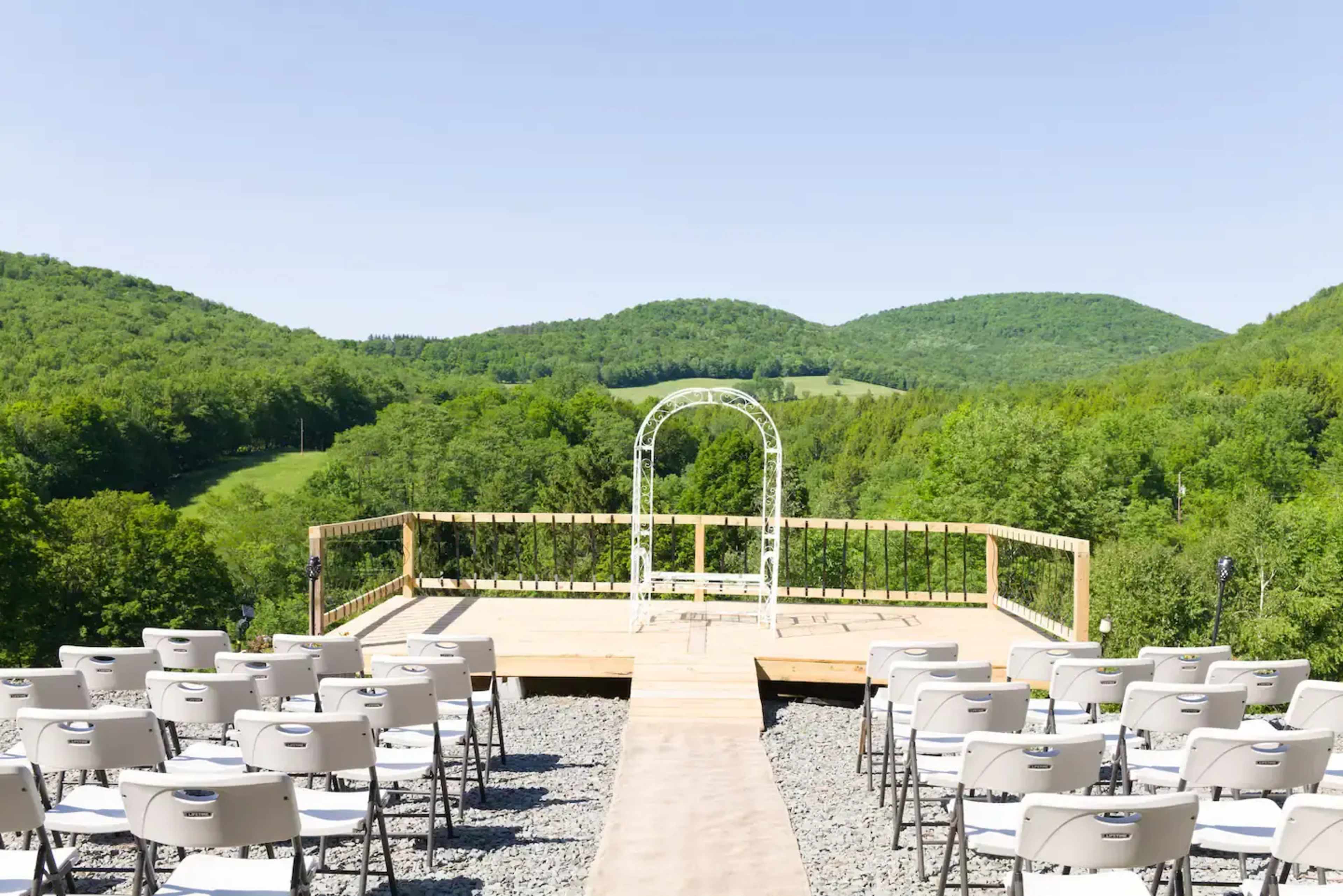 An outdoor venue features a wooden deck with a white arch at the center, surrounded by rows of folding chairs, overlooking a green mountainous landscape.