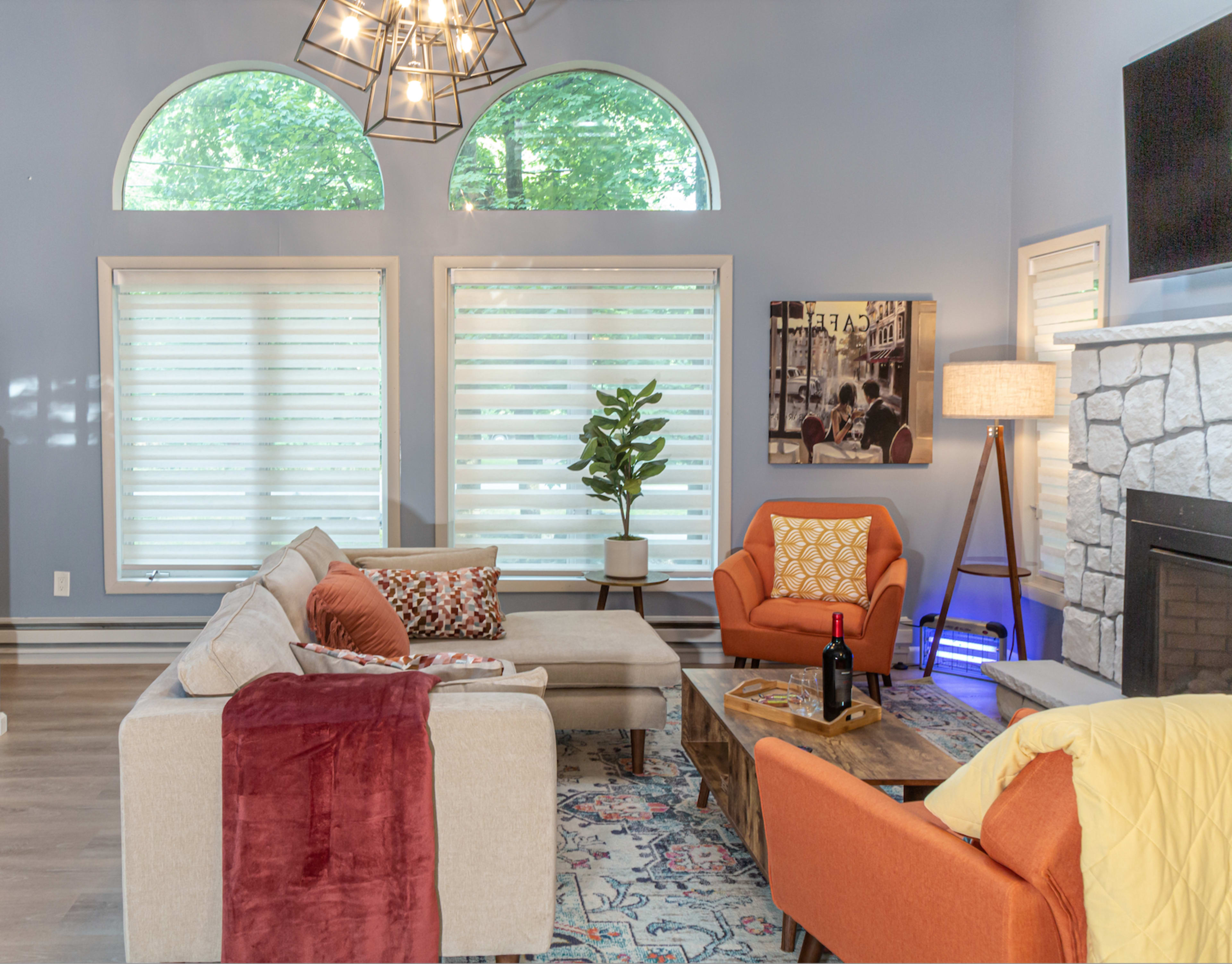 A bright living room with a sectional sofa, an orange armchair, a coffee table, and large windows with layered window treatments.