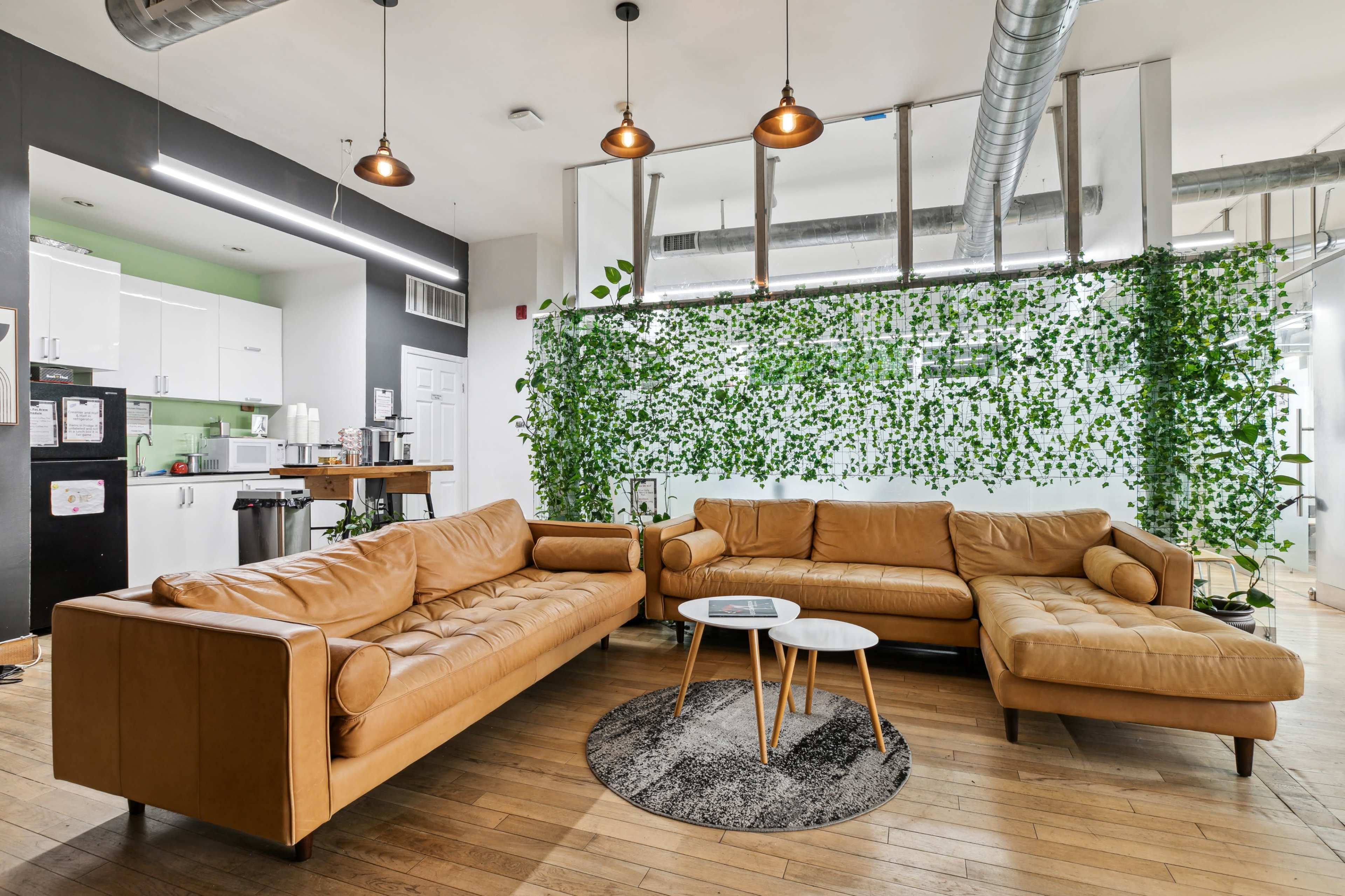 A modern interior space featuring a large, tan leather sectional sofa, a round coffee table, and a wall with green foliage, with a kitchenette visible in the background.