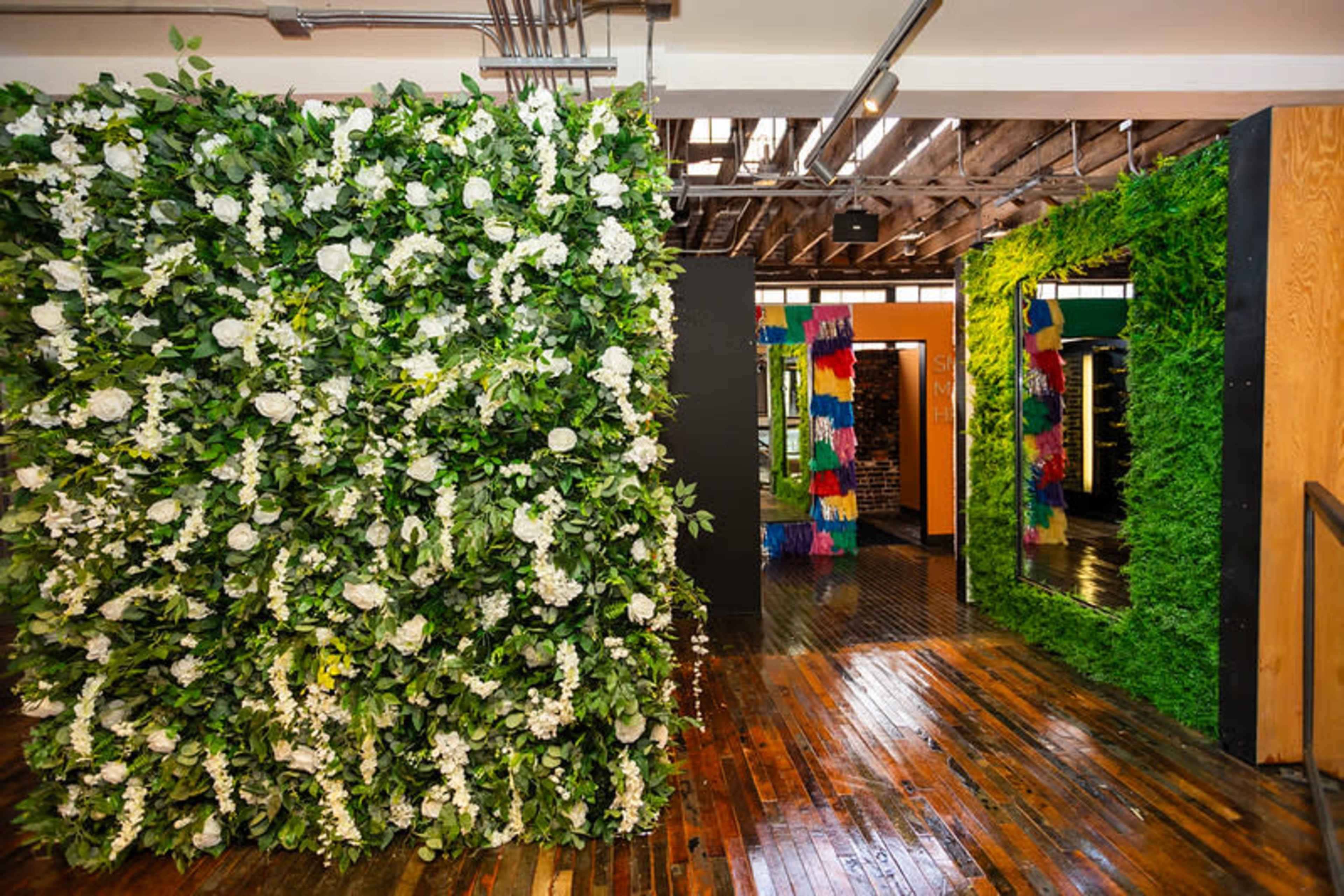 The image shows a brightly lit interior space featuring walls adorned with lush greenery and artificial flowers, alongside mirrors framed in colorful decorations.