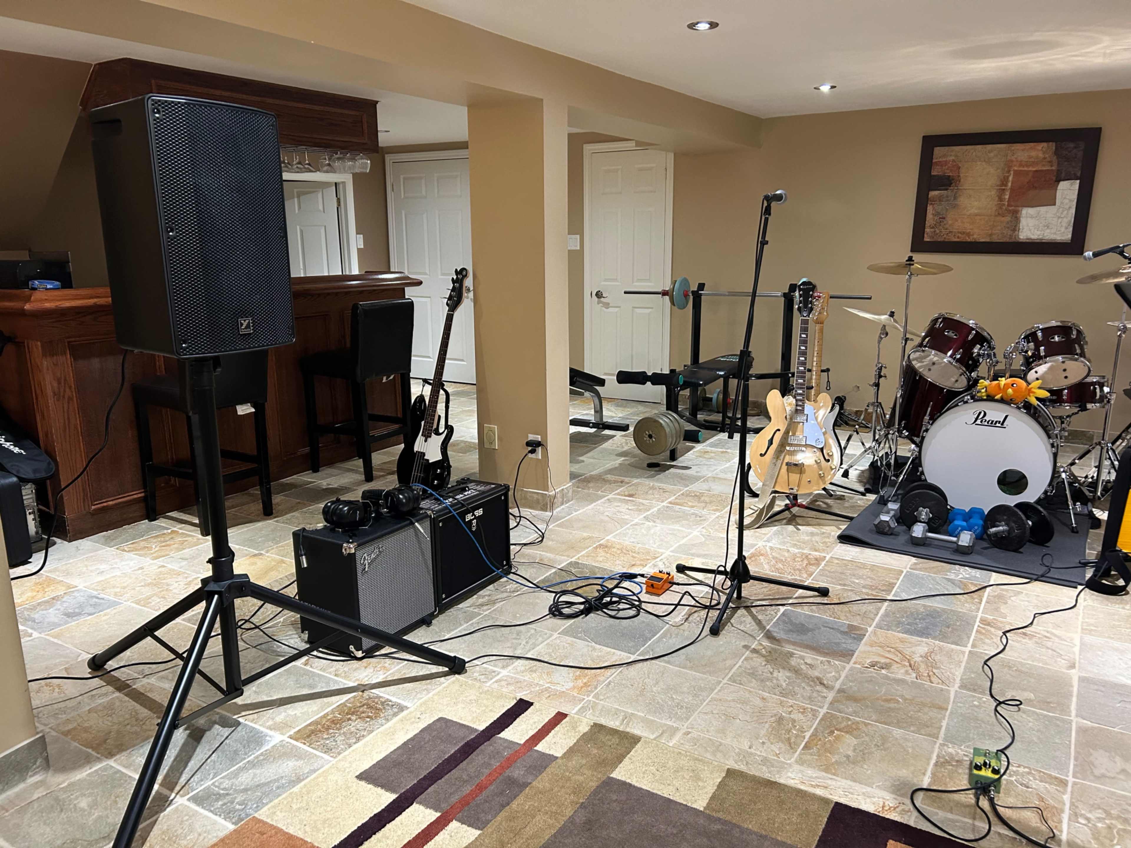 A basement music setup with amplifiers, guitars, a microphone, and a drum kit arranged around a bar area.