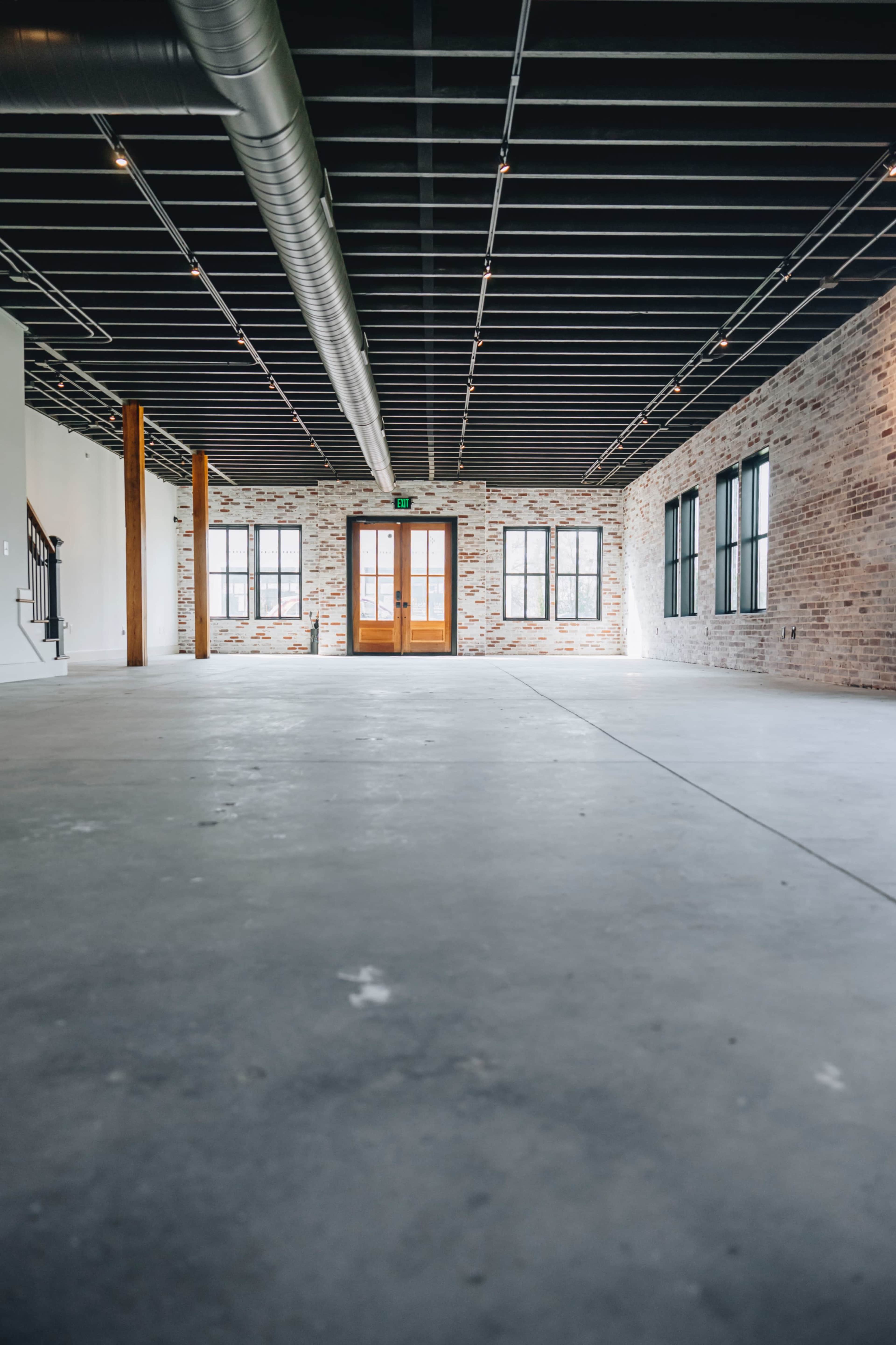 An empty room with concrete flooring, exposed brick walls, and large windows on one side, featuring a set of double doors at the entrance.