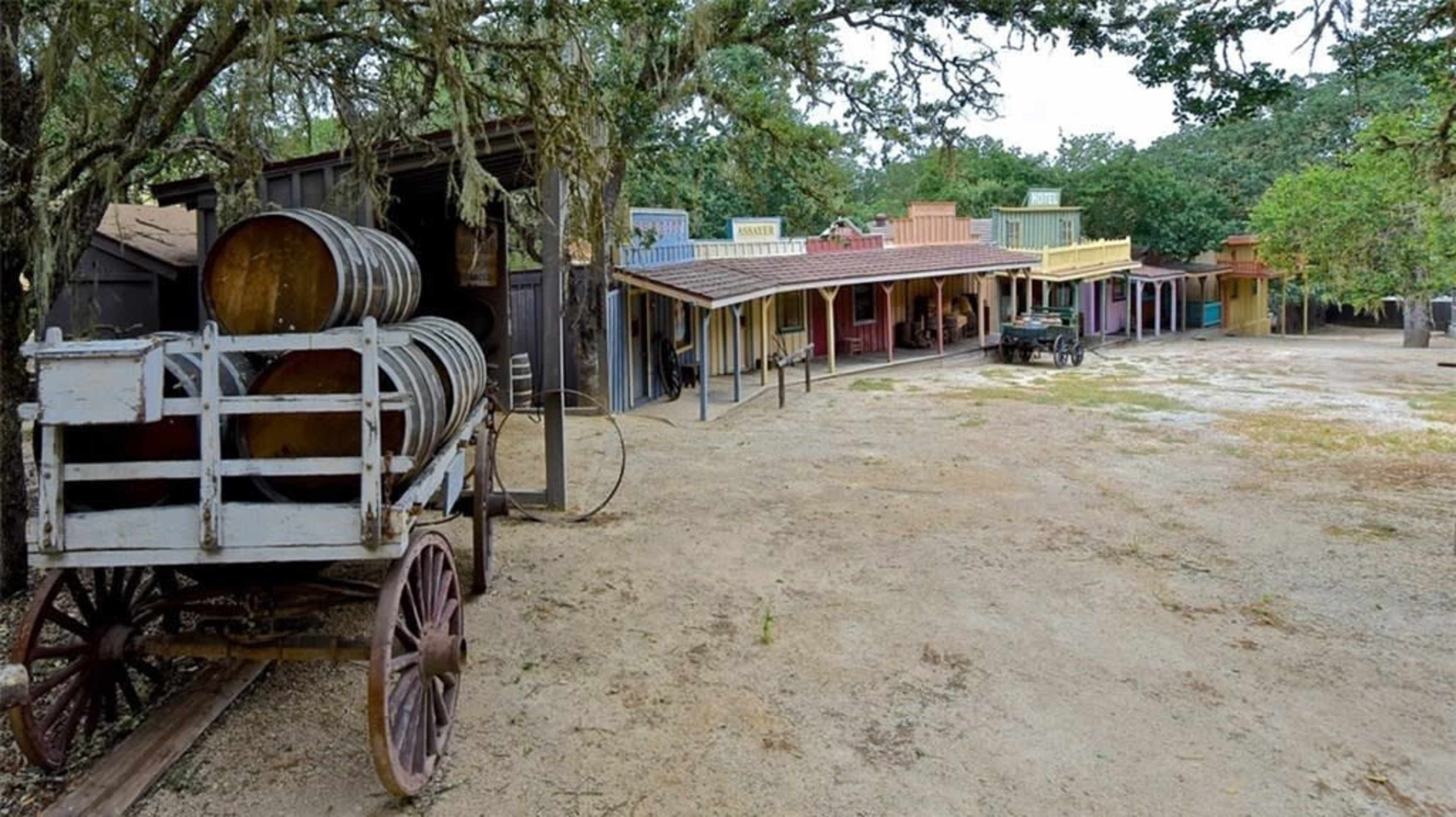 A wooden wagon with barrels is positioned near a row of colorful historic buildings under trees.