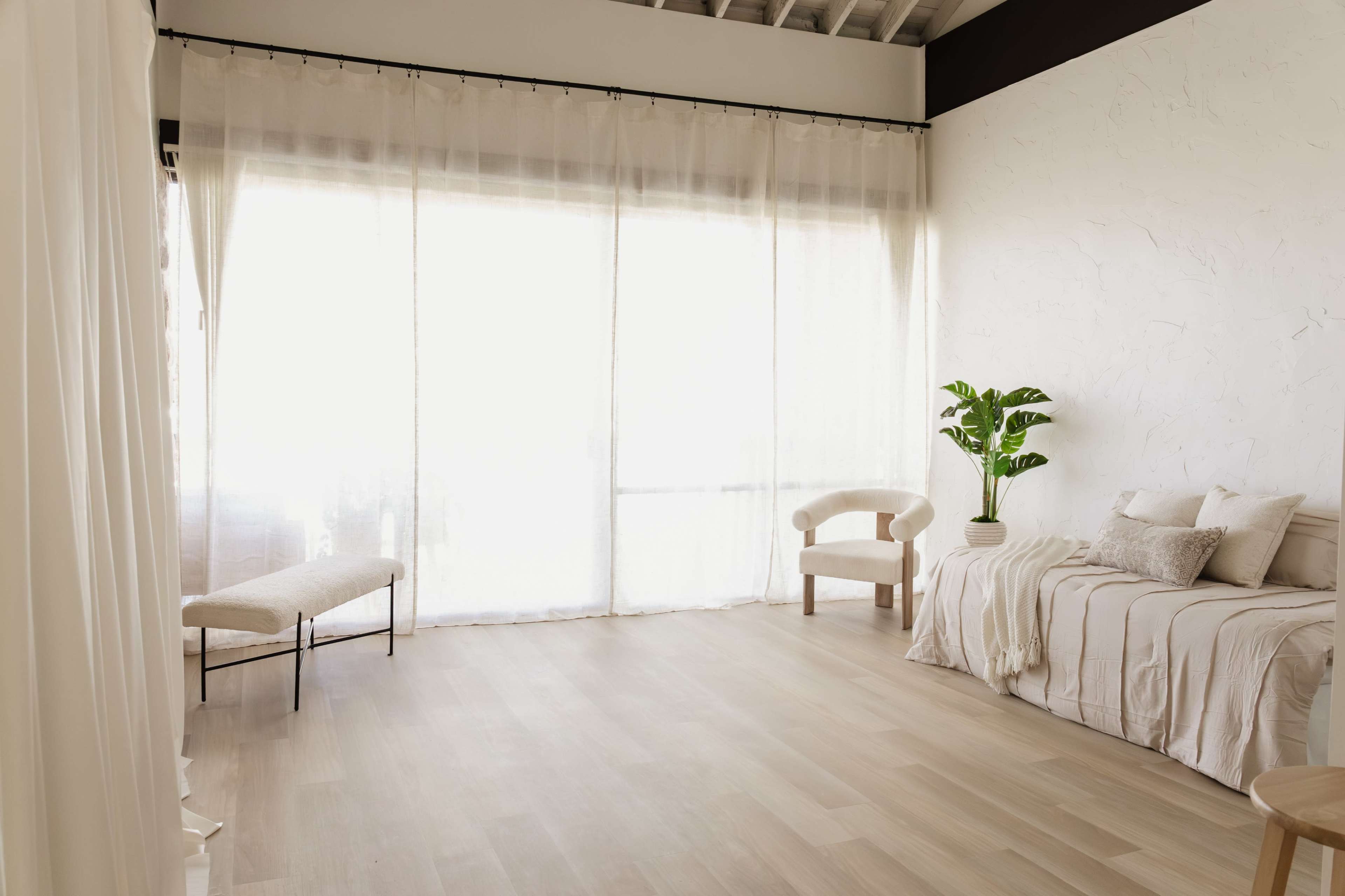 The image shows a minimalist room with large windows covered by sheer curtains, featuring a simple bench, a couch with decorative pillows, and a potted plant.