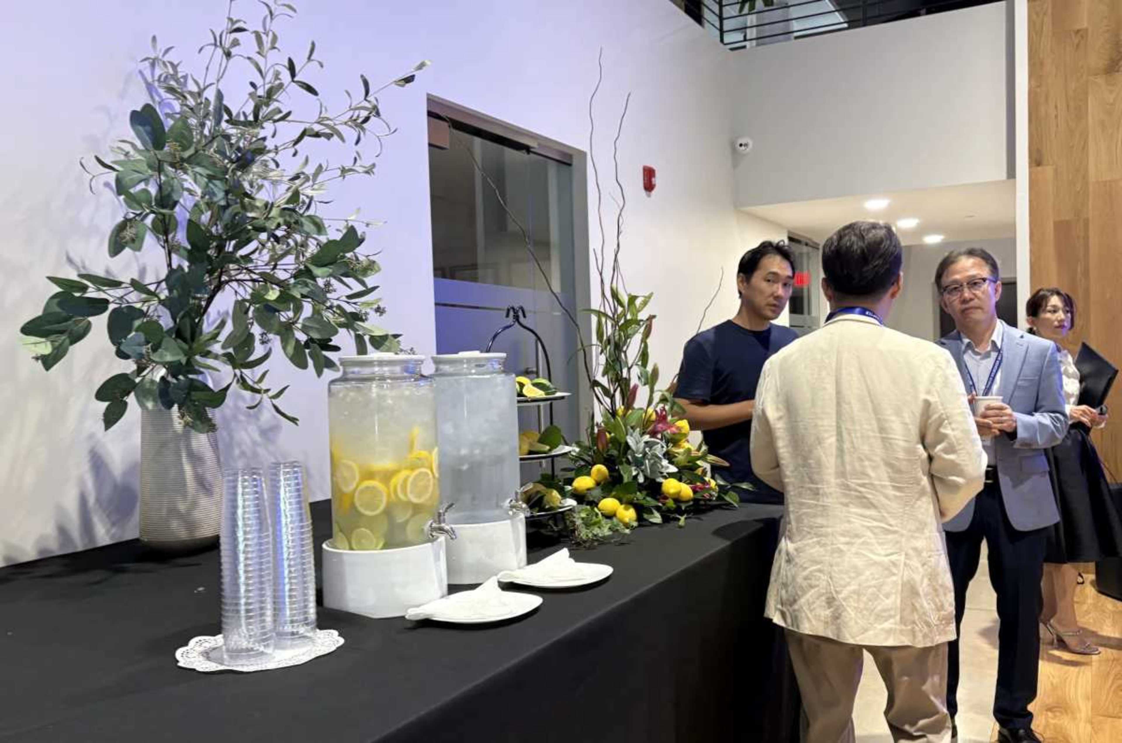 A group of individuals is engaged in conversation near a refreshment table adorned with a floral arrangement and labeled drink dispensers containing water and lemon slices.