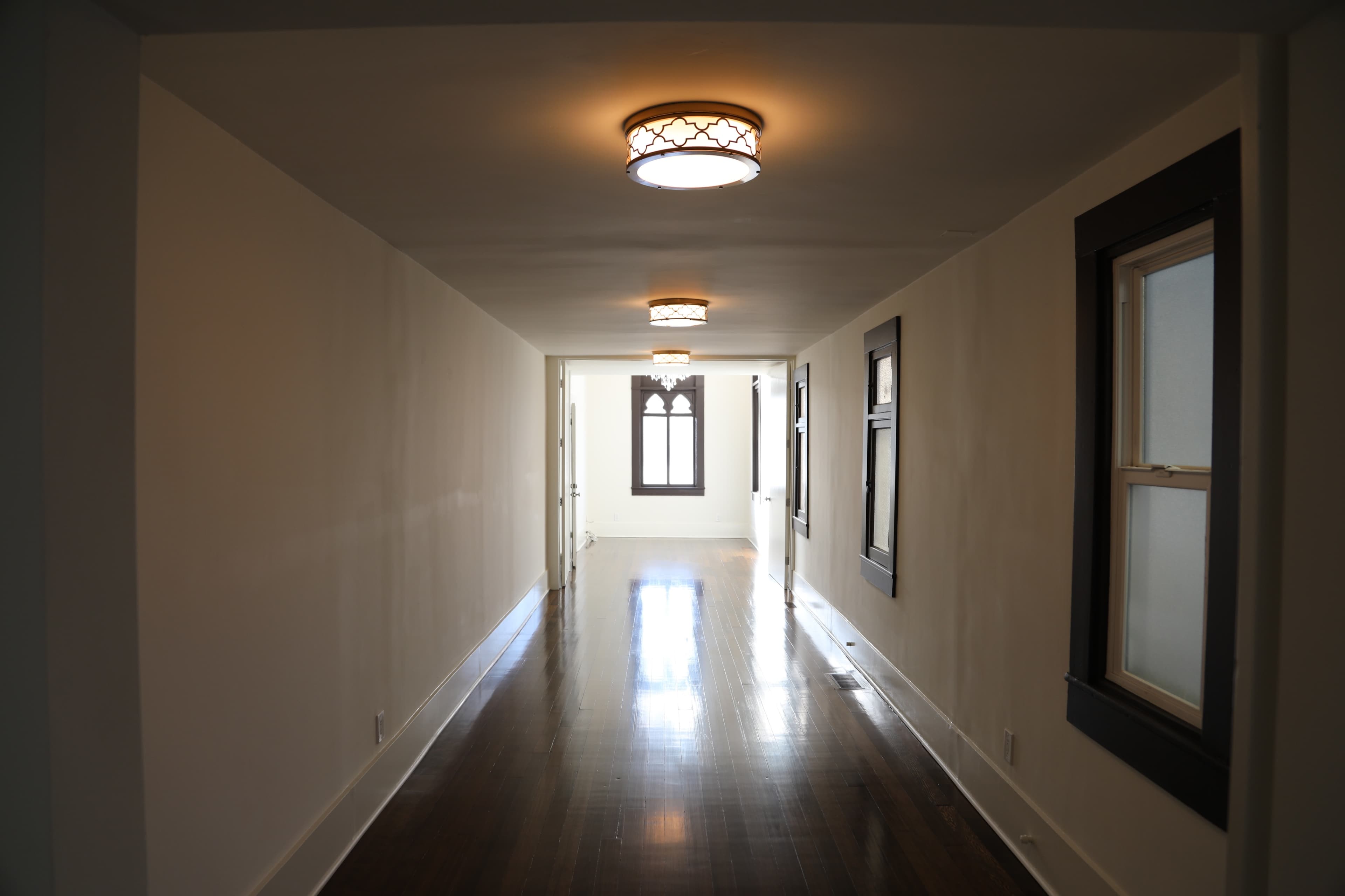 A long, narrow hallway with wooden floors, light fixtures on the ceiling, and windows along the walls.