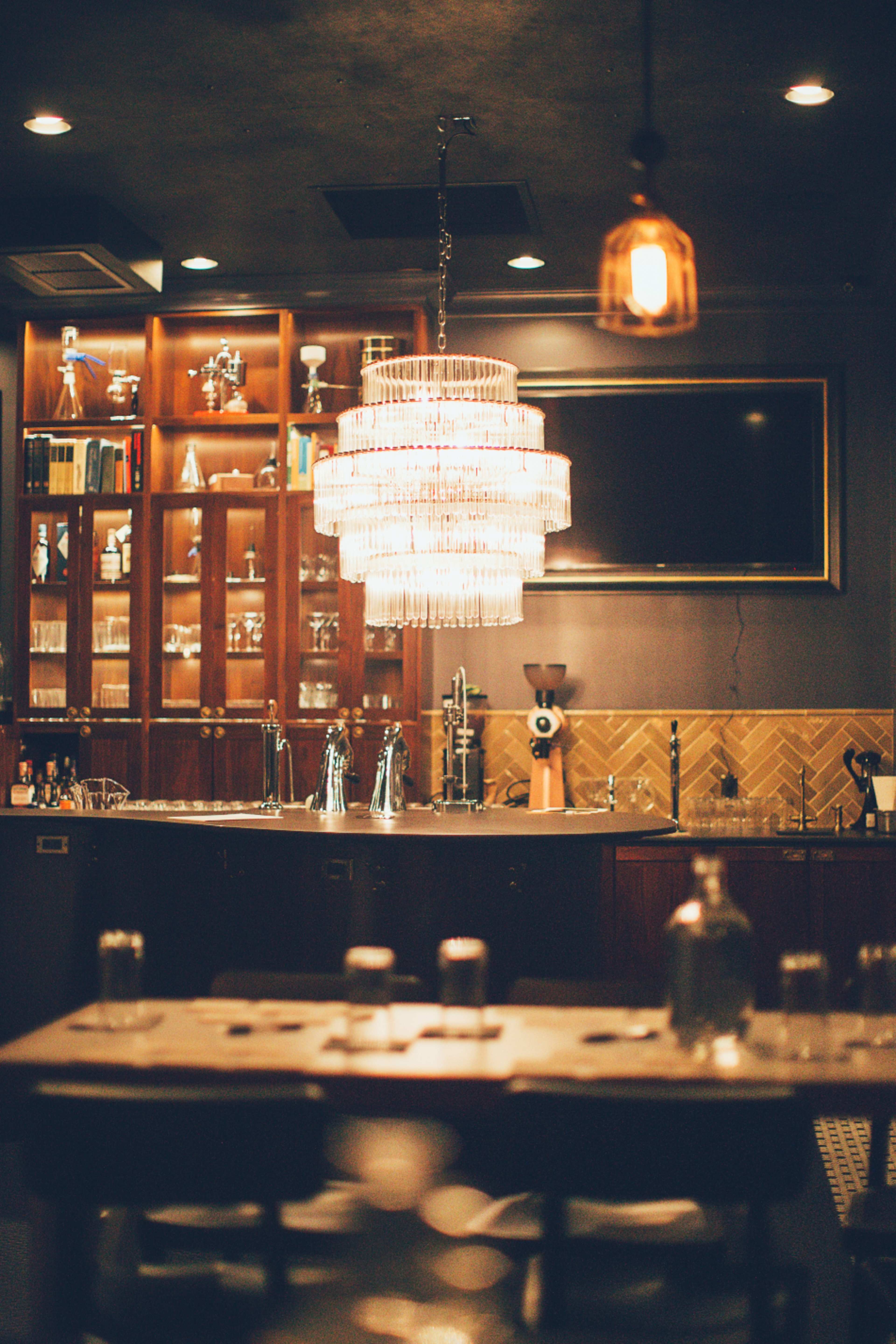 A dimly lit bar with a glass chandelier, wooden shelving filled with glassware, and a tiled counter.
