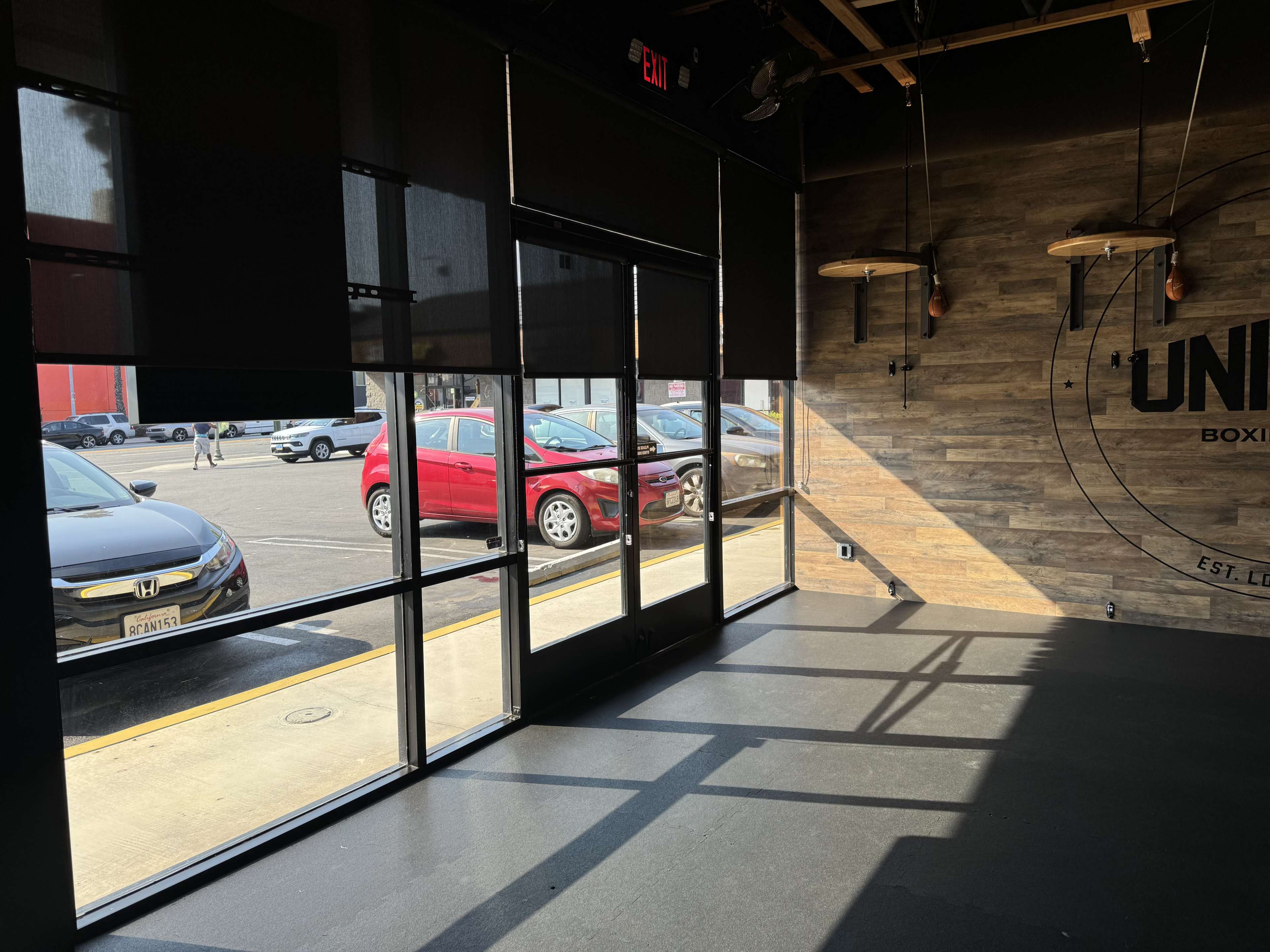 The interior of a gym features large windows allowing natural light to illuminate the space, with parked cars visible outside.
