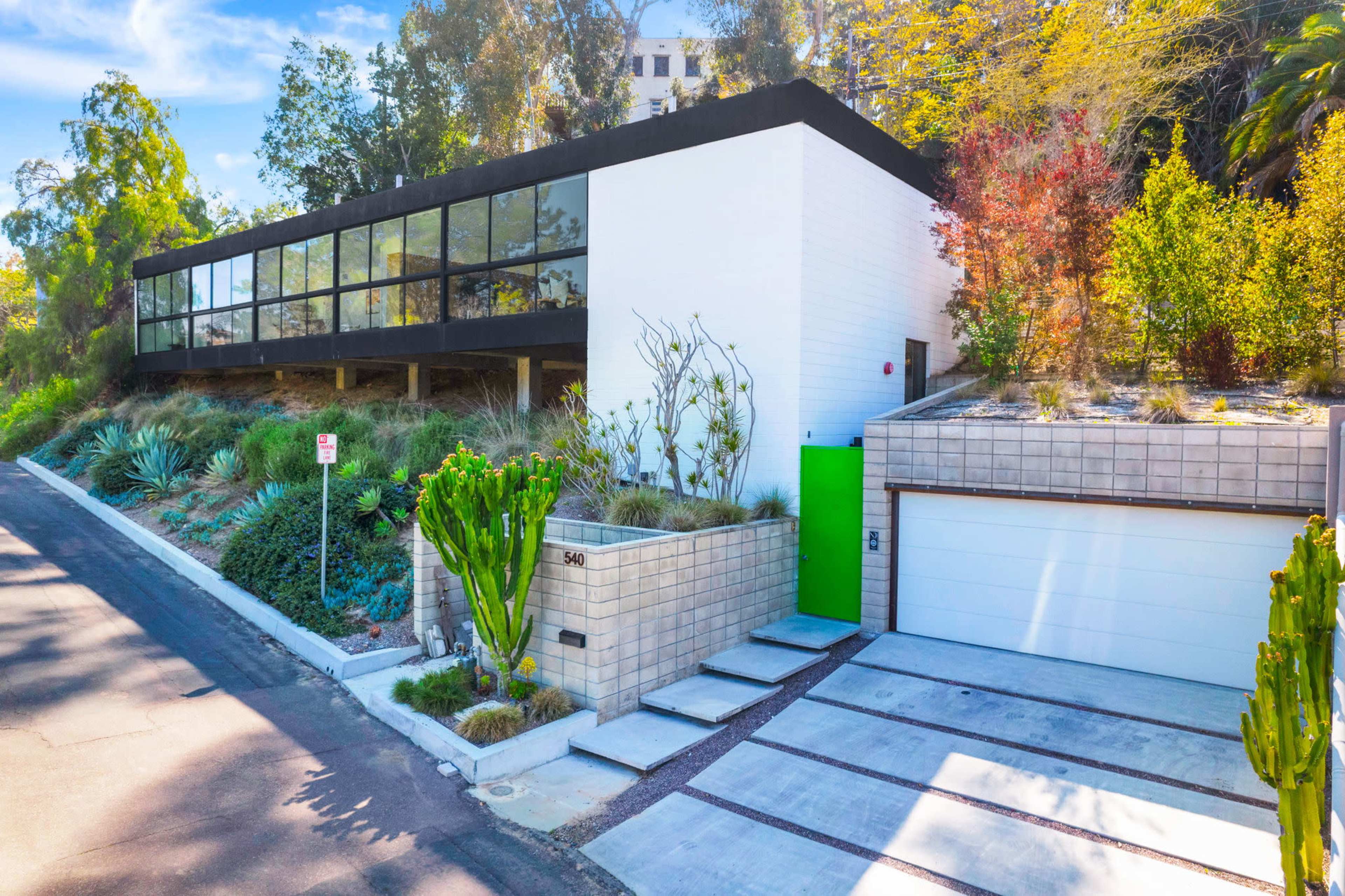 A modern house with a flat roof, large glass windows, and a bright green front door is set on a hillside surrounded by landscaped greenery and cacti.