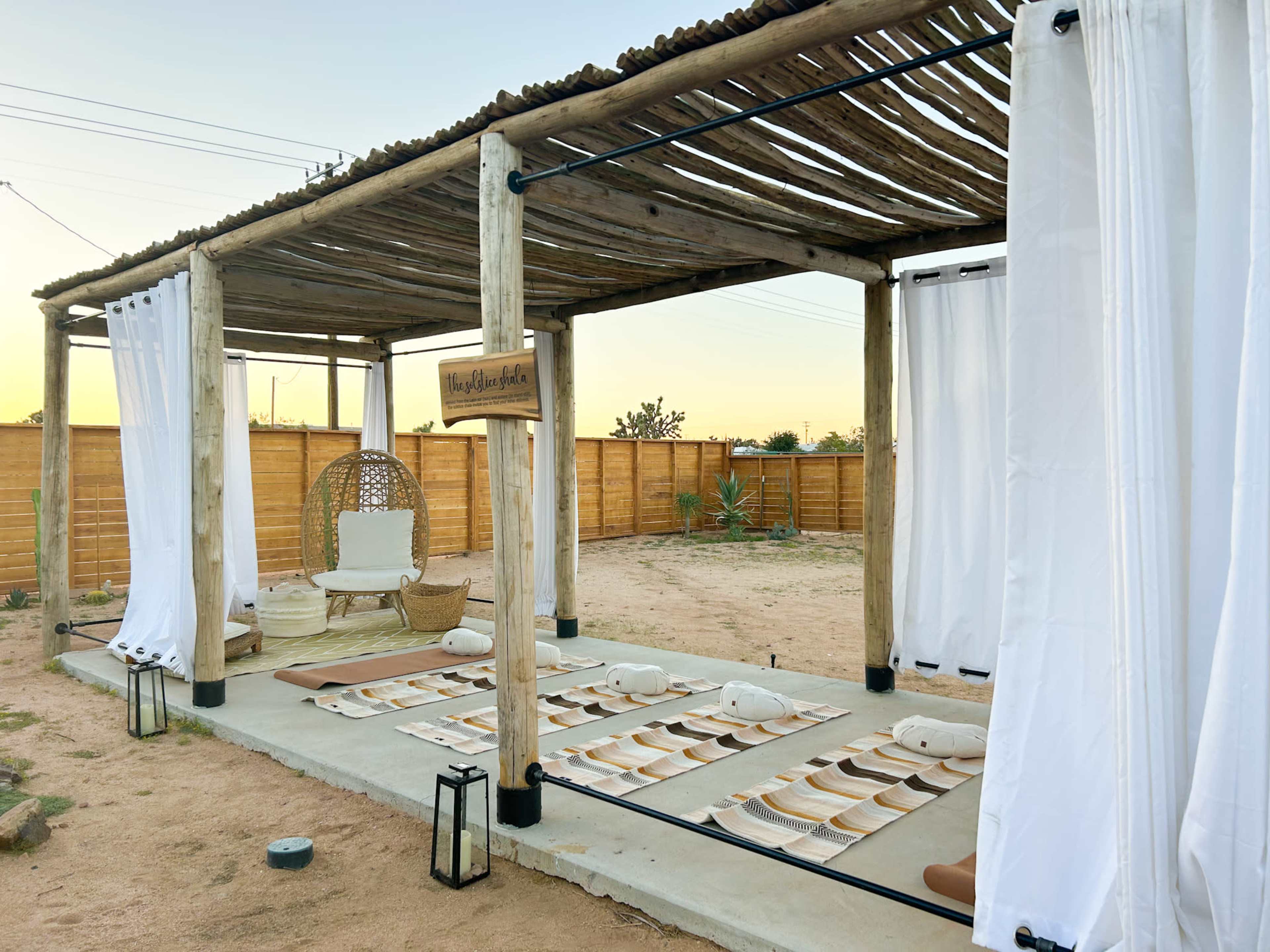A structure with a thatched roof and white curtains houses a seating area with cushions on a sandy surface, surrounded by a wooden fence and desert vegetation.