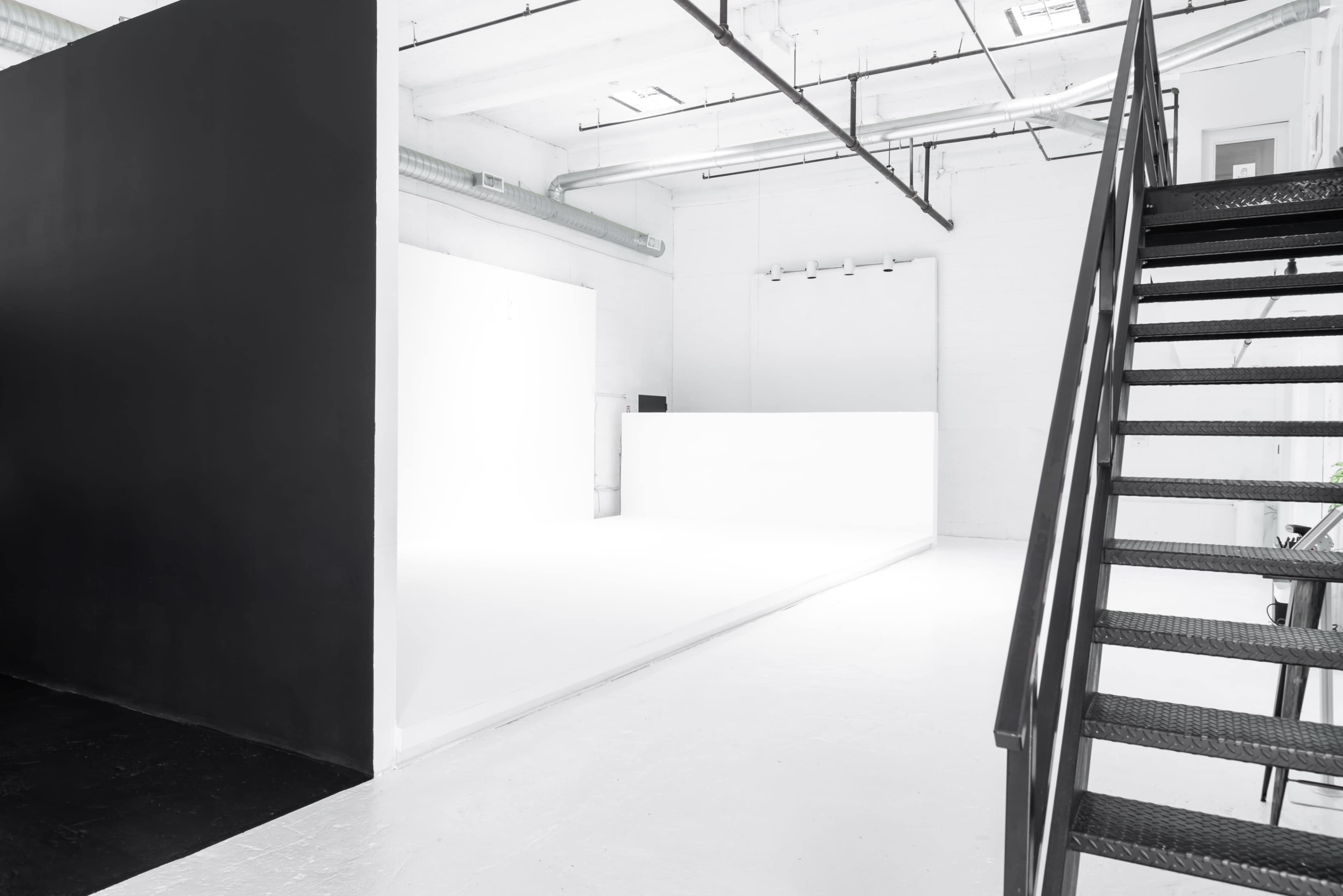 The image shows an industrial interior space featuring contrasting black and white walls, a large white platform, and a metal staircase in the foreground.