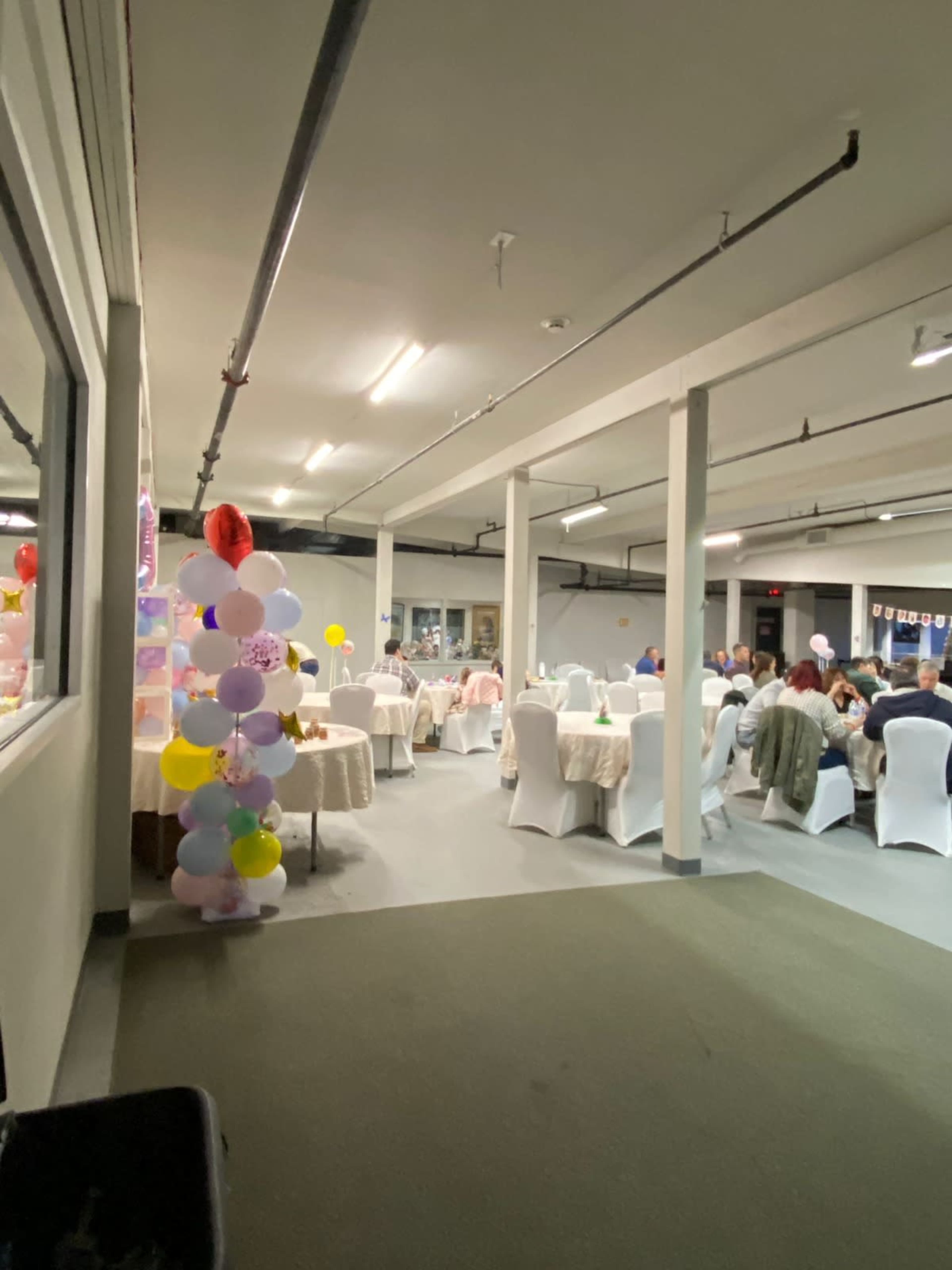 The image shows a room set up for a gathering, with tables dressed in white tablecloths and a colorful balloon column along one side.